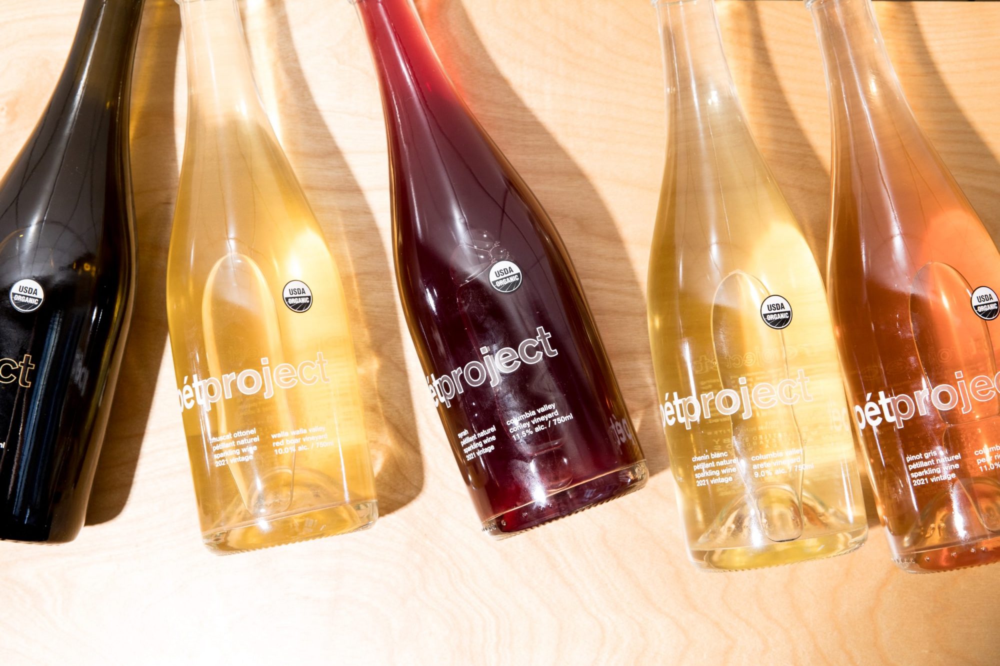 Seattle's best commercial food and hospitality photographer Five glass bottles of pét-nat wine labeled pétproject are arranged on a light wooden surface, each containing different colored wine from dark red to pale yellow and orange—captured by Seattles best commercial food lifestyle photographer. captured by seattle's best food and lifestyle photographer Brooke Fitts