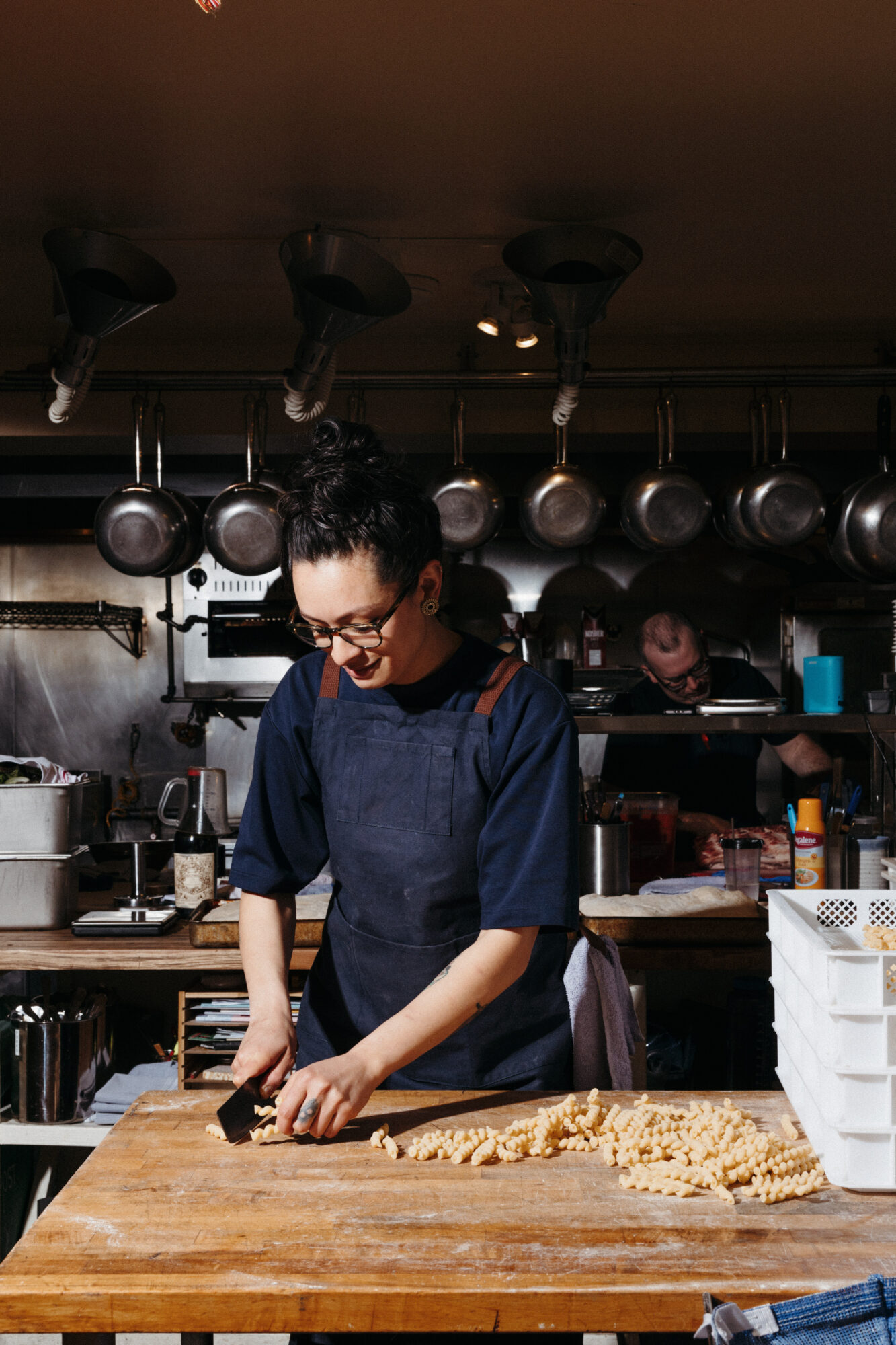 Seattle's best commercial food and hospitality photographer A woman with glasses and an apron smiles while cutting fresh pasta on a wooden table in a busy kitchen—captured by Seattles best commercial food lifestyle photographer, with pots and pans hanging in the background. captured by seattle's best food and lifestyle photographer Brooke Fitts