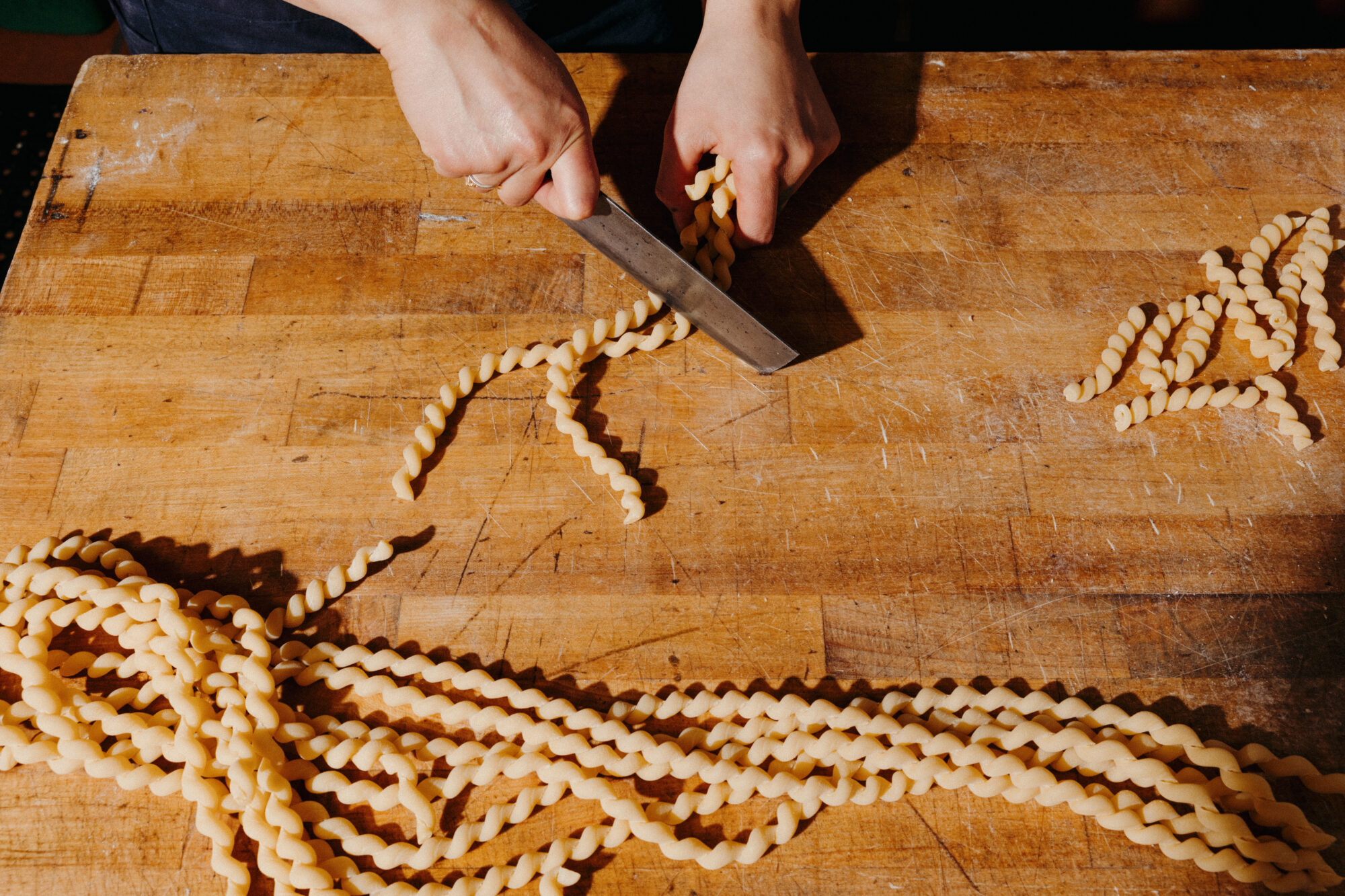 Seattle's best commercial food and hospitality photographer Hands cutting twisted pasta dough with a large knife on a wooden surface, several pieces of uncooked pasta scattered around—captured by Seattles best commercial food lifestyle photographer. captured by seattle's best food and lifestyle photographer Brooke Fitts