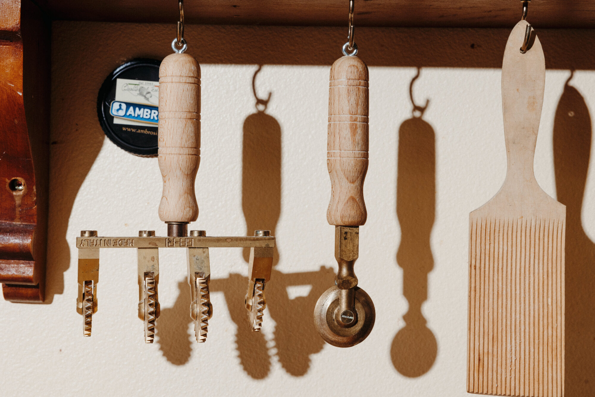 Seattle's best commercial food and hospitality photographer Wooden-handled pasta cutting tools and a gnocchi board hang on hooks against a beige wall, casting distinct shadows—captured by Seattles best commercial food lifestyle photographer. A round black-topped container rests in the background. captured by seattle's best food and lifestyle photographer Brooke Fitts