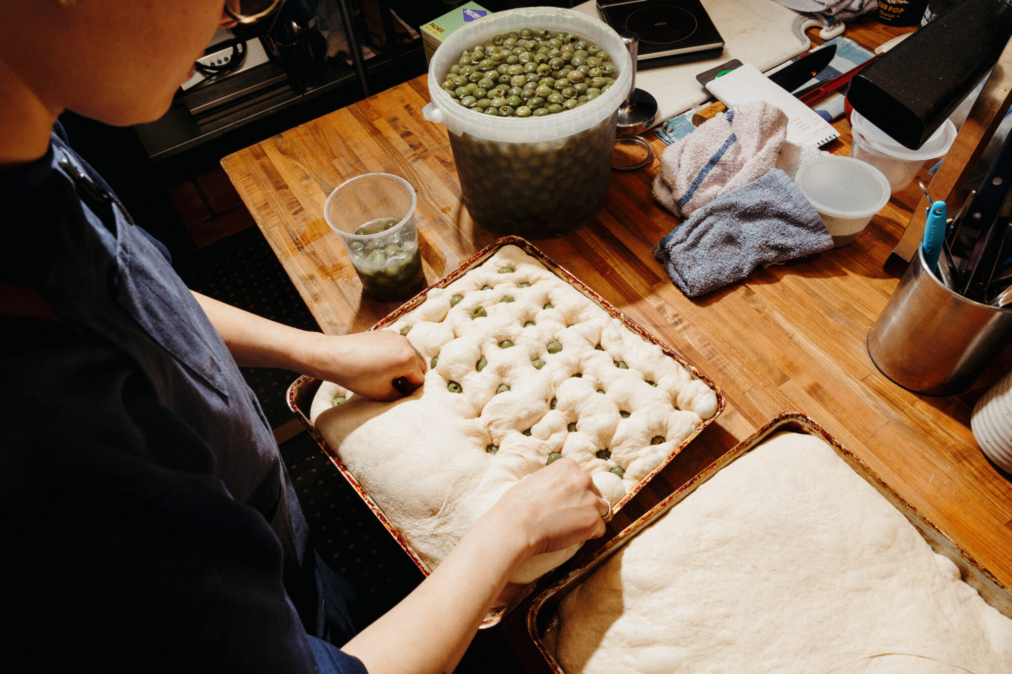 Seattle's best commercial food and hospitality photographer A person presses green olives into dough on a tray, preparing focaccia. Another tray with plain dough, a large container of olives, and kitchen utensils sit on the wooden countertop—captured by Seattles best commercial food lifestyle photographer. captured by seattle's best food and lifestyle photographer Brooke Fitts