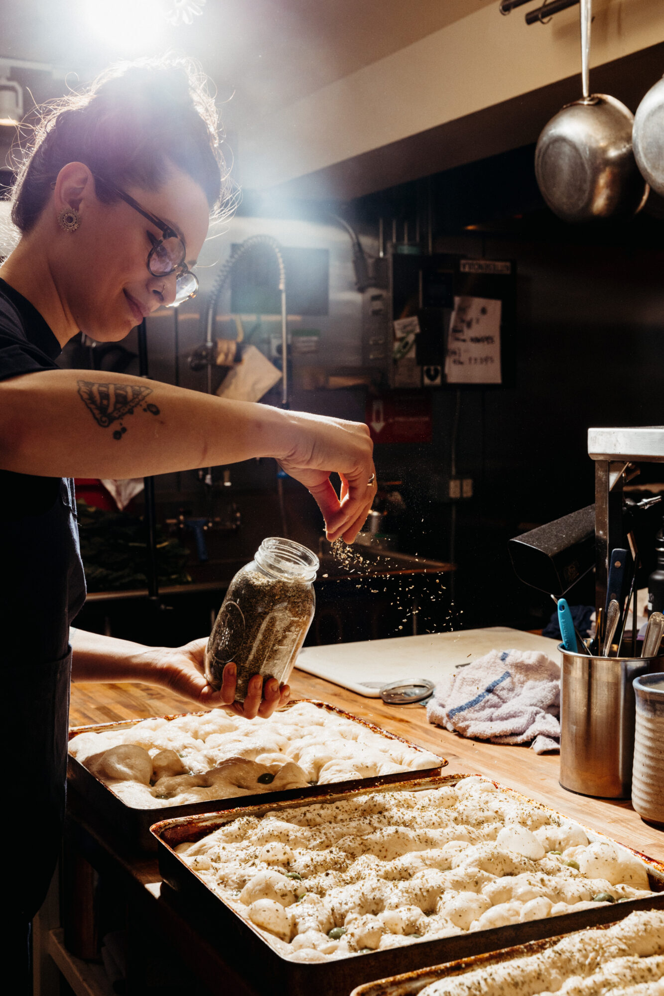 Seattle's best commercial food and hospitality photographer A woman with glasses sprinkles herbs from a jar onto trays of dough in a sunlit kitchen—an inviting scene captured by Seattles best commercial food lifestyle photographer. captured by seattle's best food and lifestyle photographer Brooke Fitts