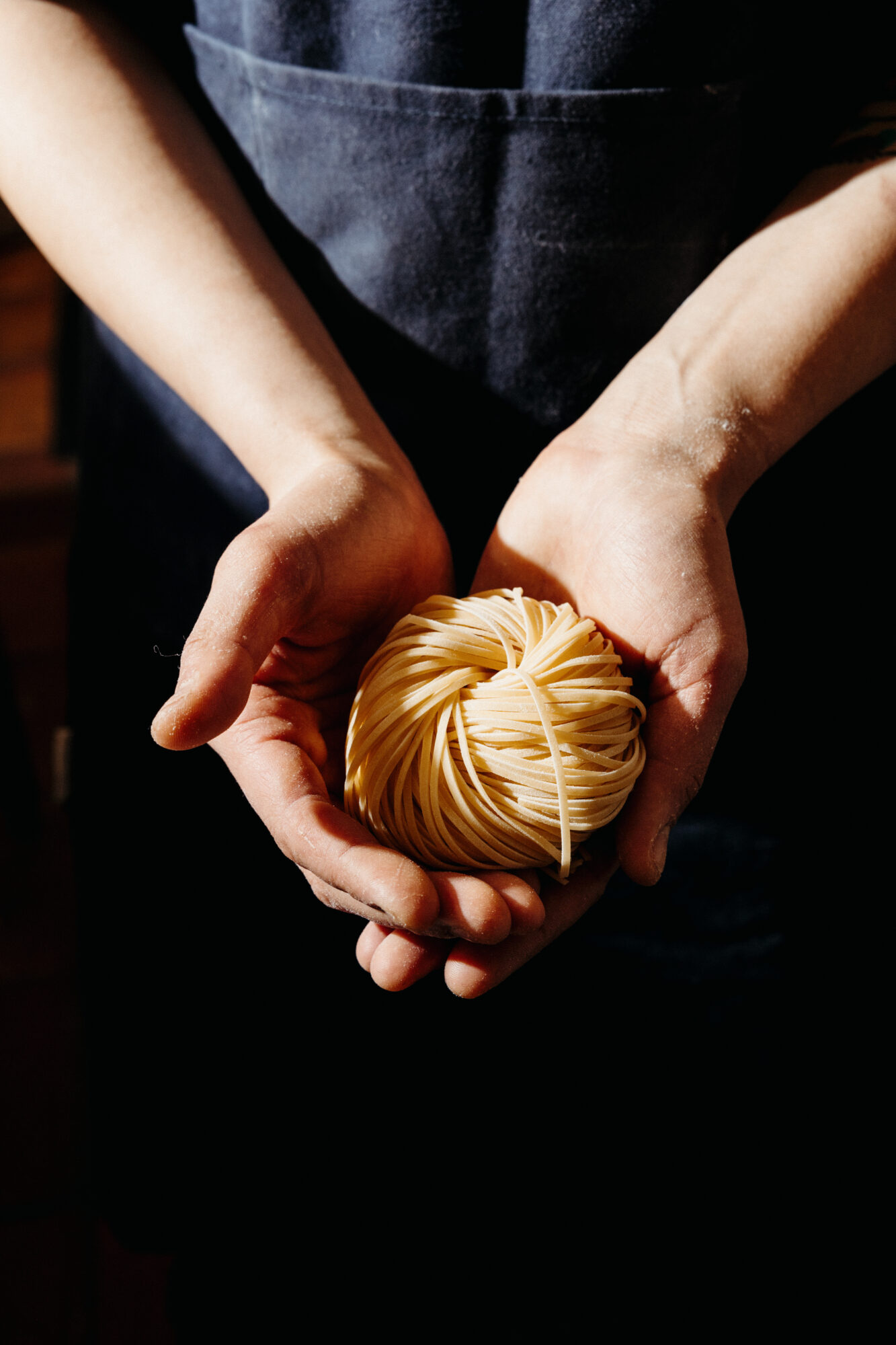 Seattle's best commercial food and hospitality photographer A person in a dark apron holds a neatly coiled nest of uncooked pasta in their cupped hands, illuminated by natural light against a dark background—captured by Seattles best commercial food lifestyle photographer. captured by seattle's best food and lifestyle photographer Brooke Fitts