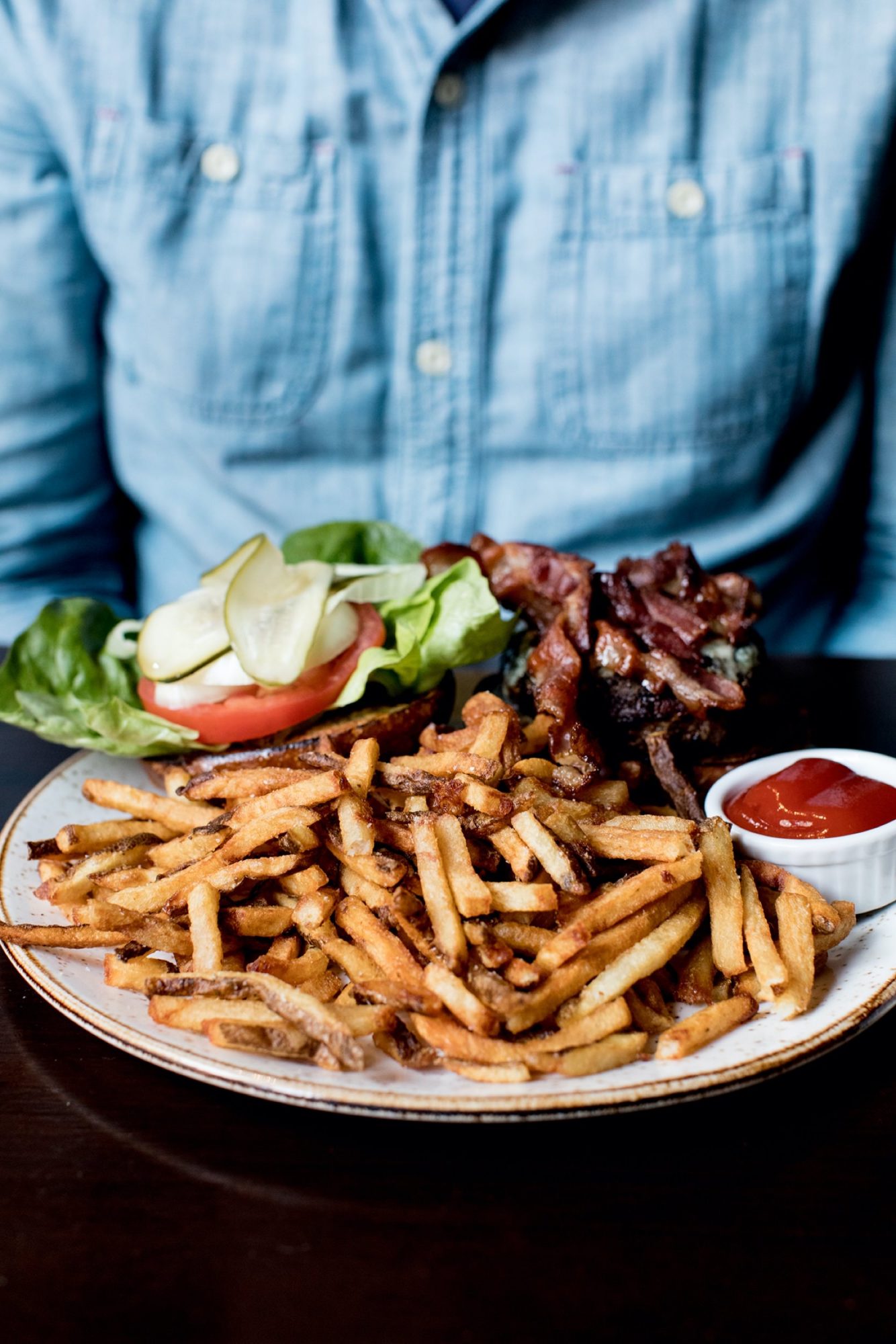 Seattle's best commercial food and hospitality photographer A person in a blue denim shirt sits at a table with French fries, a bacon cheeseburger piled with lettuce, tomato, and pickles, and a small dish of ketchup—captured by Seattles best commercial food lifestyle photographer. captured by seattle's best food and lifestyle photographer Brooke Fitts