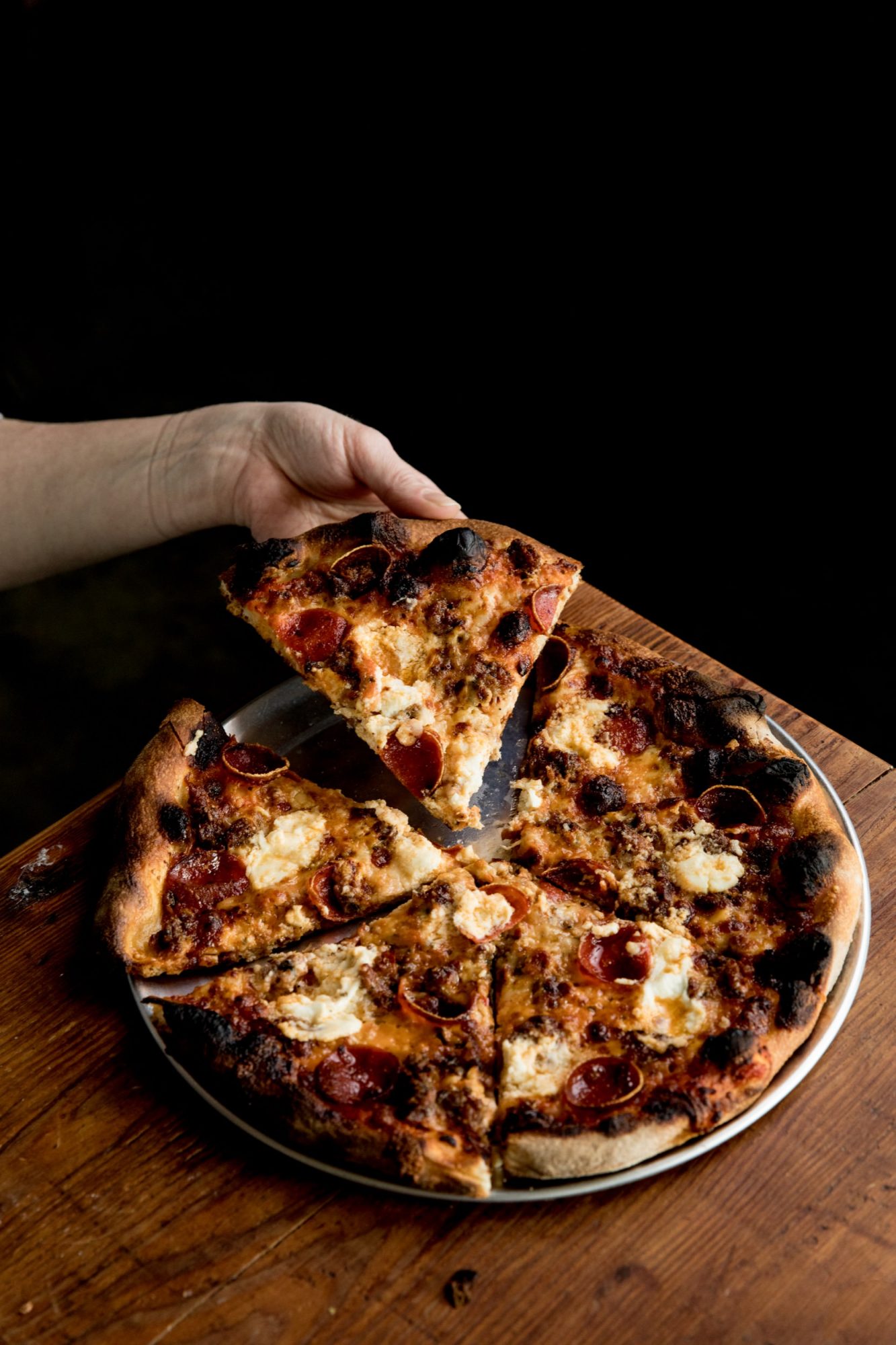 Seattle's best commercial food and hospitality photographer A hand lifts a slice of pepperoni pizza with melted cheese from a metal tray on a wooden table, perfectly captured by Seattles best commercial food lifestyle photographer. The pizza features a charred, crispy crust and is cut into six slices. captured by seattle's best food and lifestyle photographer Brooke Fitts