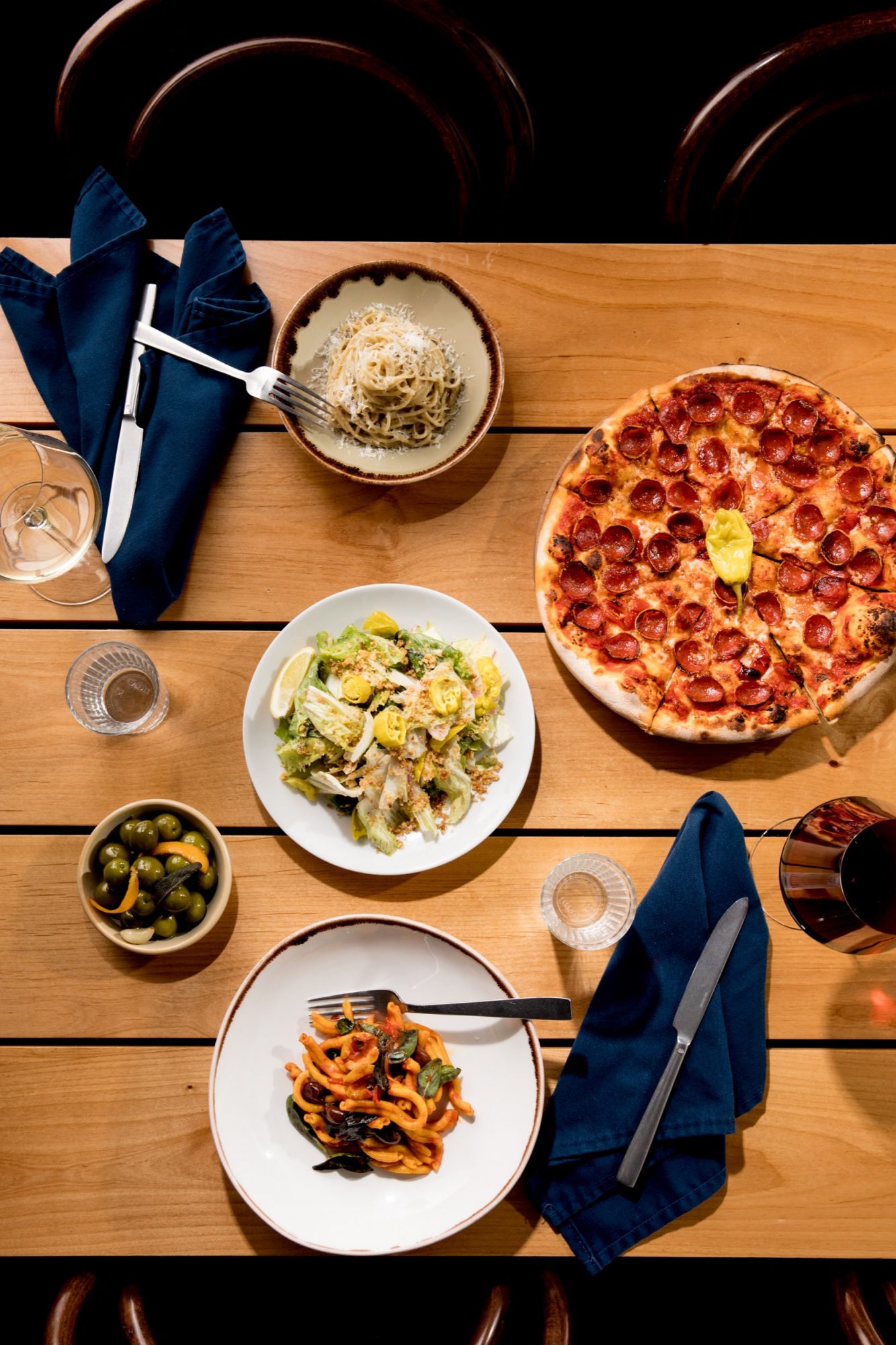 Seattle's best commercial food and hospitality photographer Overhead view of a wooden table set with a pepperoni pizza, pasta, Caesar salad, marinated olives, red wine, and blue napkins with cutlery—captured by Seattles best commercial food lifestyle photographer. captured by seattle's best food and lifestyle photographer Brooke Fitts