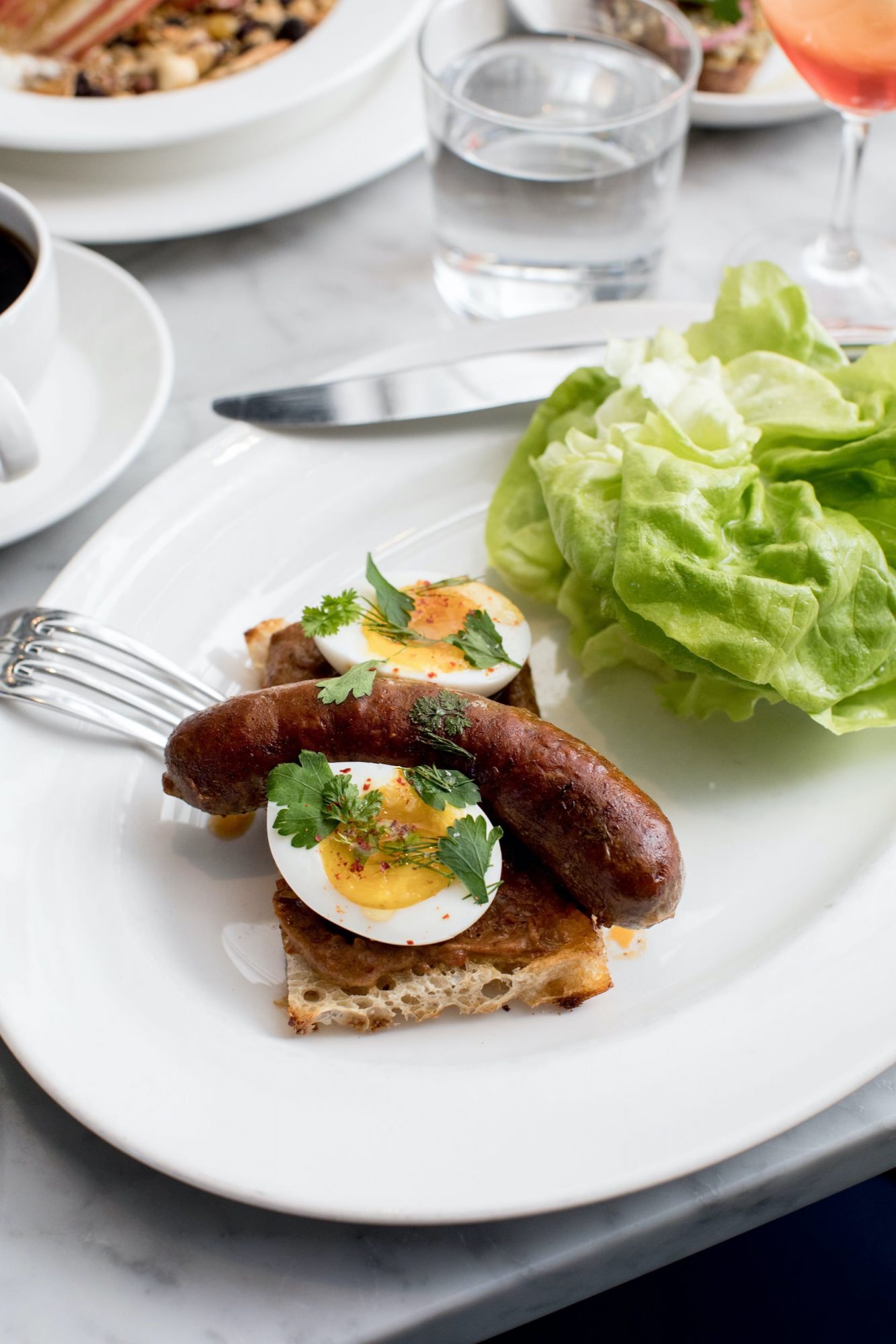 Seattle's best commercial food and hospitality photographer A plate styled by Seattles best commercial food lifestyle photographer features sausage, halved boiled eggs on toast with herbs, fresh lettuce, and a fork resting on the bread—set alongside a glass of water and other dishes in the background. captured by seattle's best food and lifestyle photographer Brooke Fitts