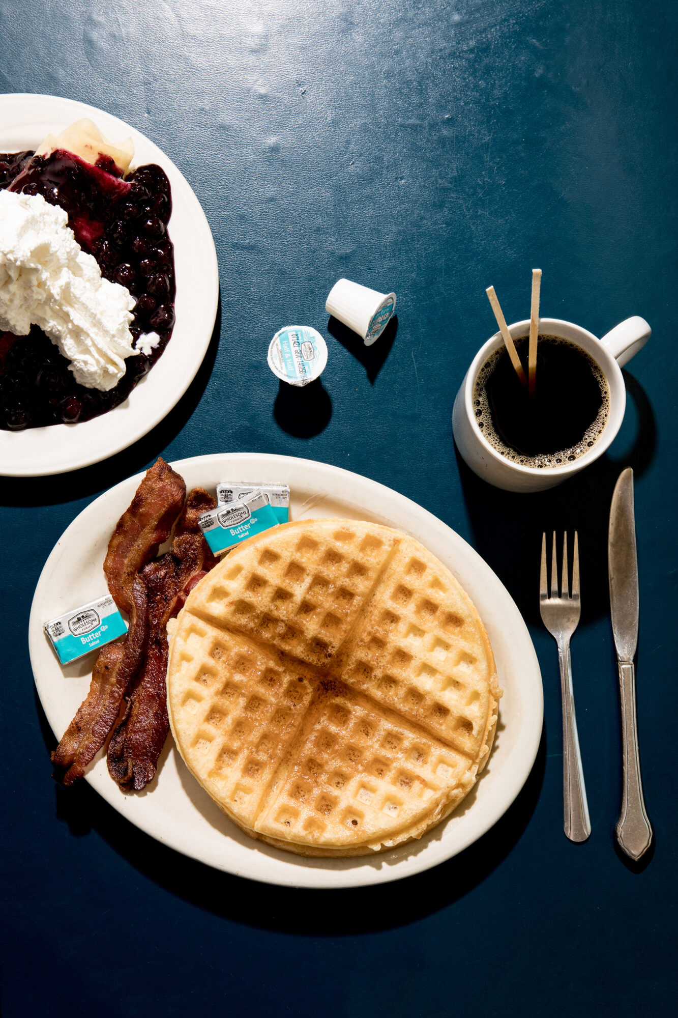 Seattle's best commercial food and hospitality photographer A breakfast of waffles with butter, strips of bacon, coffee with creamer, and utensils sits on a dark blue table—captured by Seattles best commercial food lifestyle photographer. A plate of whipped cream and berries is also partially visible. captured by seattle's best food and lifestyle photographer Brooke Fitts