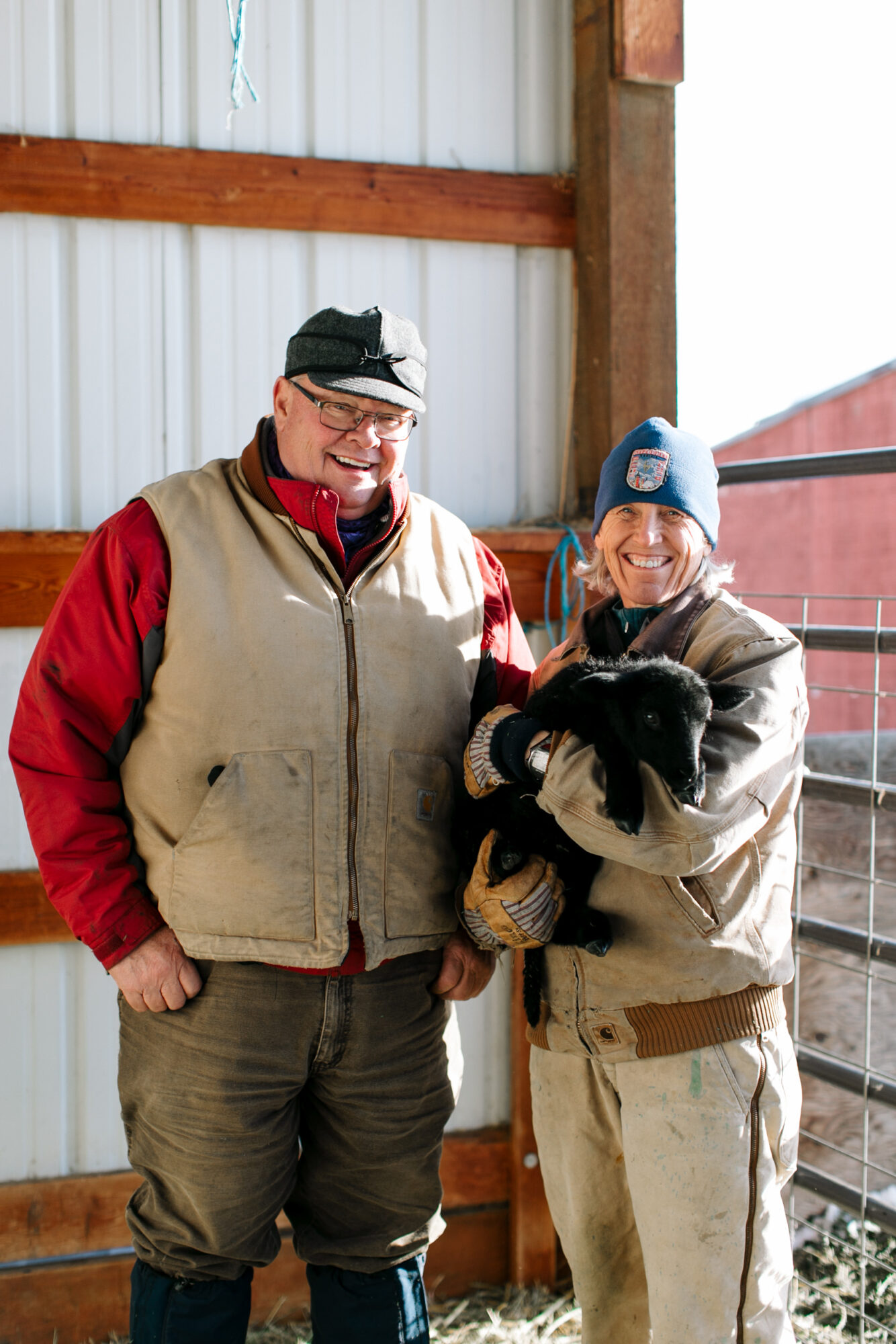 Seattle's best commercial food and hospitality photographer Two people in warm work clothes stand in a barn. One, holding a small black goat, smiles at the camera while the other stands beside them. Sunlight shines in from the side—captured by Seattles best commercial food lifestyle photographer. captured by seattle's best food and lifestyle photographer Brooke Fitts
