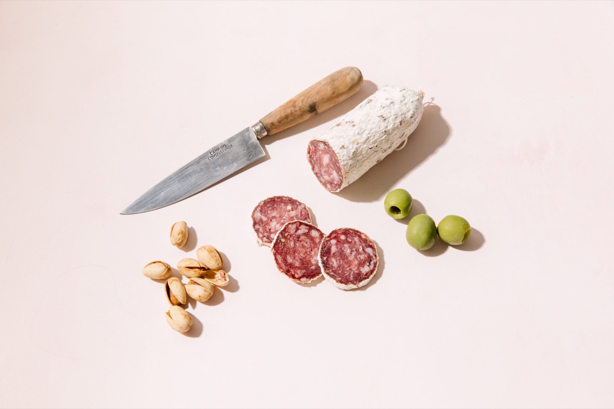 Seattle's best commercial food and hospitality photographer A knife, three green olives, a small pile of pistachios, and a partially sliced salami are artfully arranged on a light pink surface by Seattles best commercial food lifestyle photographer. captured by seattle's best food and lifestyle photographer Brooke Fitts