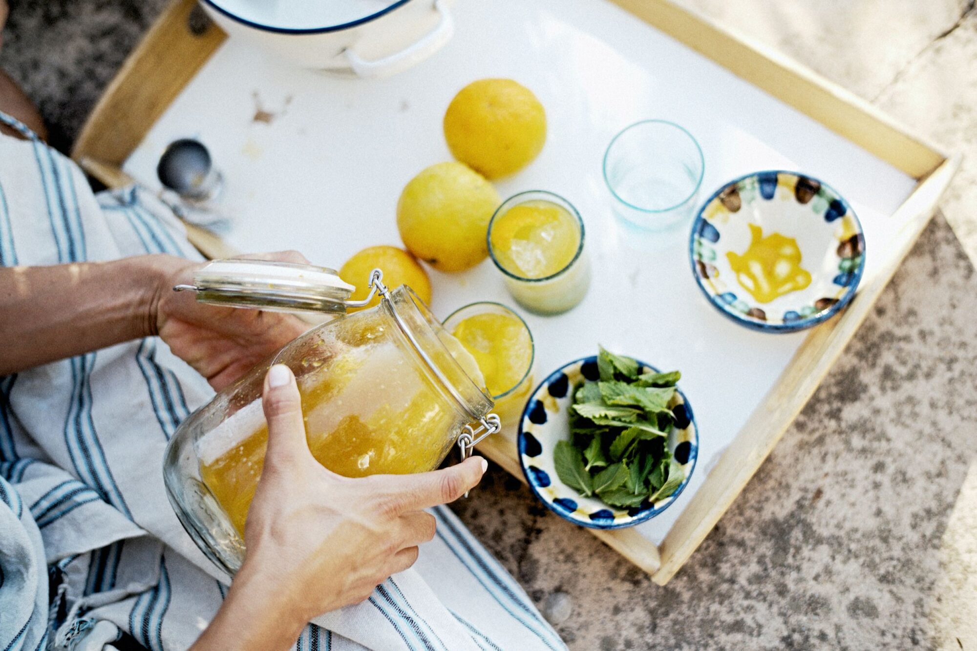 Seattle's best commercial food and hospitality photographer A person prepares a refreshing drink, holding a glass jar of yellow liquid near a tray with lemons, glasses, small bowls, and fresh mint leaves—captured by Seattles best commercial food lifestyle photographer on a sunny outdoor surface. captured by seattle's best food and lifestyle photographer Brooke Fitts