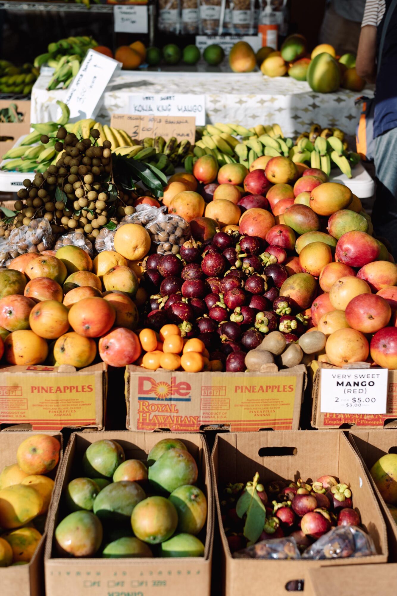 Seattle's best commercial food and hospitality photographer A colorful market stand displays various tropical fruits, including mangoes, bananas, mangosteen, pineapples, and longans, arranged in cardboard boxes under bright sunlight—captured by Seattles best commercial food lifestyle photographer. captured by seattle's best food and lifestyle photographer Brooke Fitts