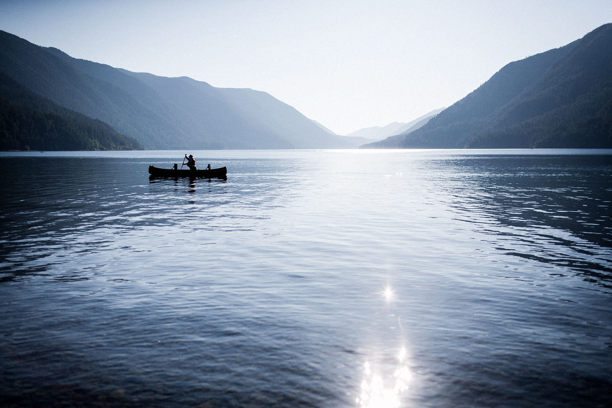 Seattle's best commercial food and hospitality photographer A person paddles a canoe on a calm lake surrounded by mountains under a clear sky, with sunlight reflecting off the water—captured beautifully by Seattles best commercial food lifestyle photographer. captured by seattle's best food and lifestyle photographer Brooke Fitts