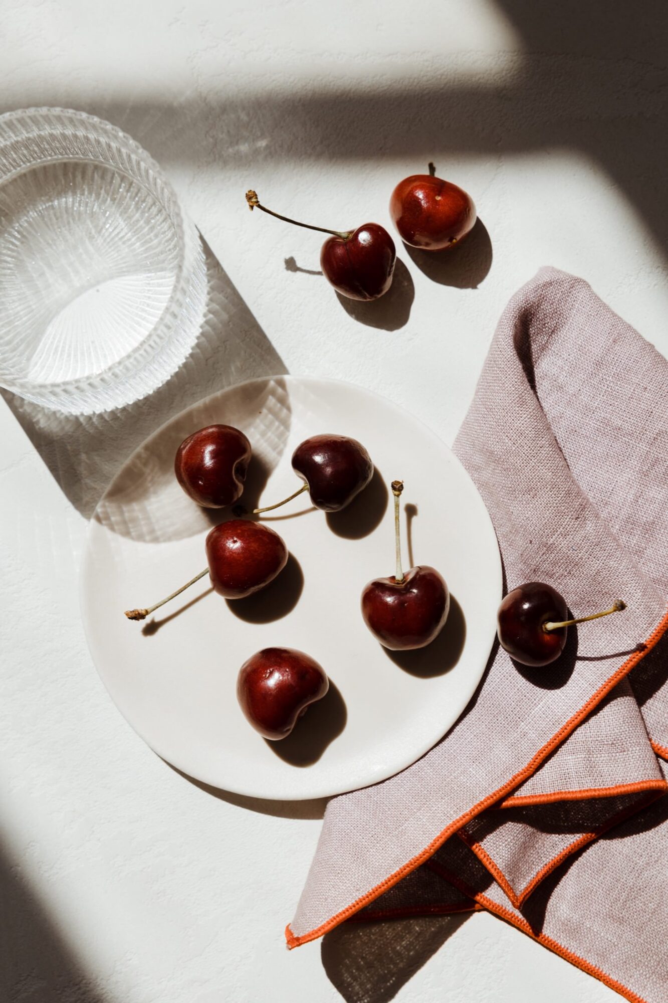 Seattle's best commercial food and hospitality photographer A white plate with six cherries sits on a light surface beside a pink napkin with orange trim; three more cherries and a clear glass cup are nearby, styled by Seattles best commercial food lifestyle photographer in soft sunlight and shadow. captured by seattle's best food and lifestyle photographer Brooke Fitts
