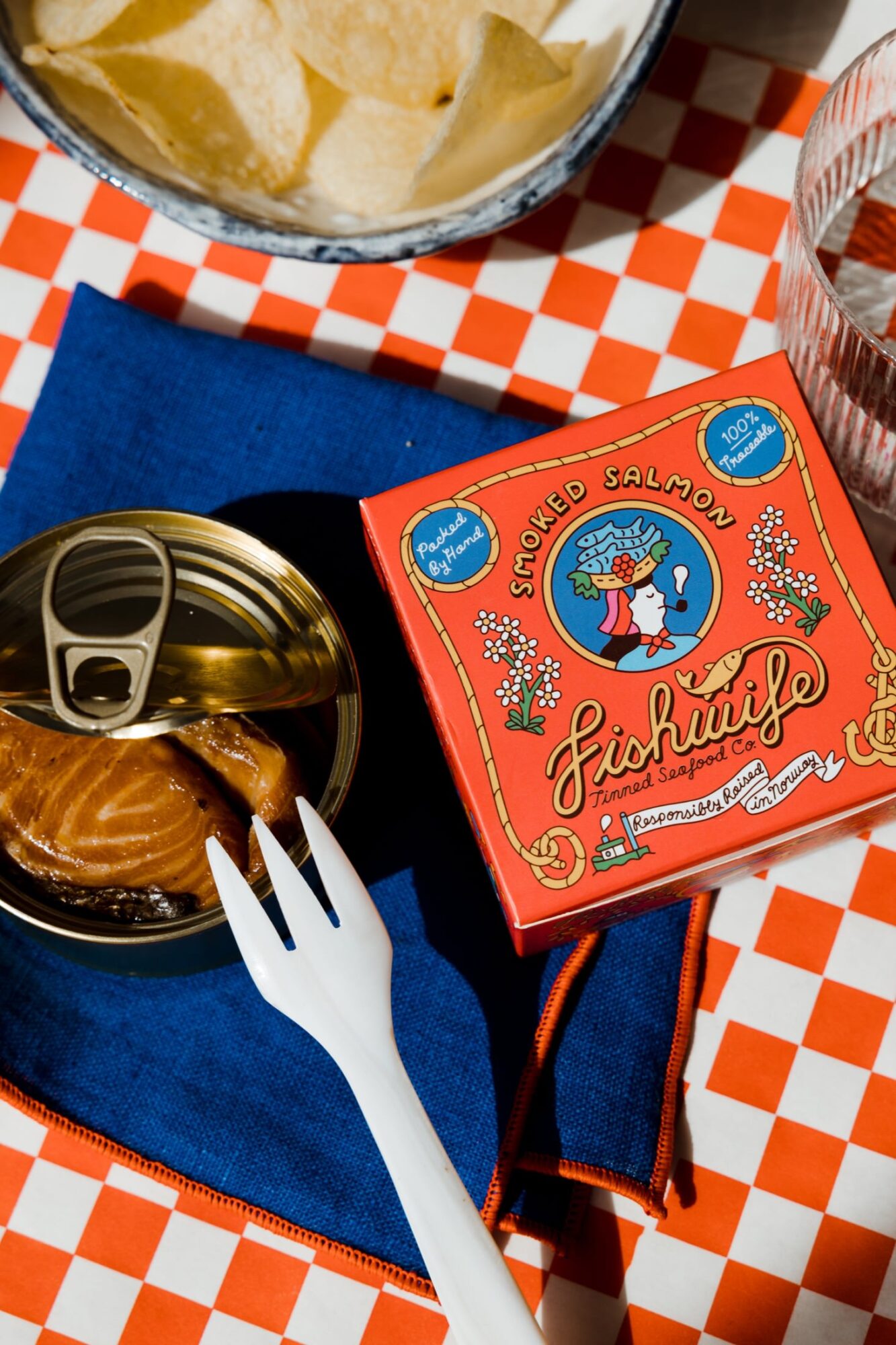 Seattle's best commercial food and hospitality photographer A can of smoked salmon, partially opened, sits on a blue napkin with a plastic fork beside it. Captured by Seattles best commercial food lifestyle photographer, the scene includes a colorful Fishwife box, chips, and water on a red checkered tablecloth. captured by seattle's best food and lifestyle photographer Brooke Fitts