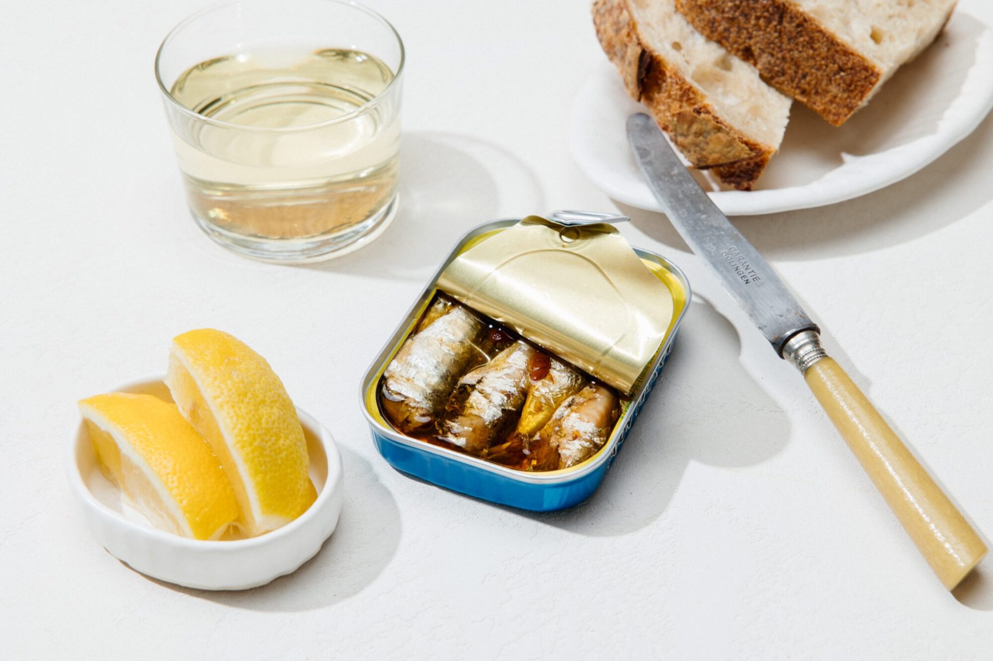 Seattle's best commercial food and hospitality photographer A tin of sardines with the lid partially opened sits on a white surface next to lemon wedges, a glass of white wine, slices of bread on a plate, and a knife—captured by Seattles best commercial food lifestyle photographer. captured by seattle's best food and lifestyle photographer Brooke Fitts