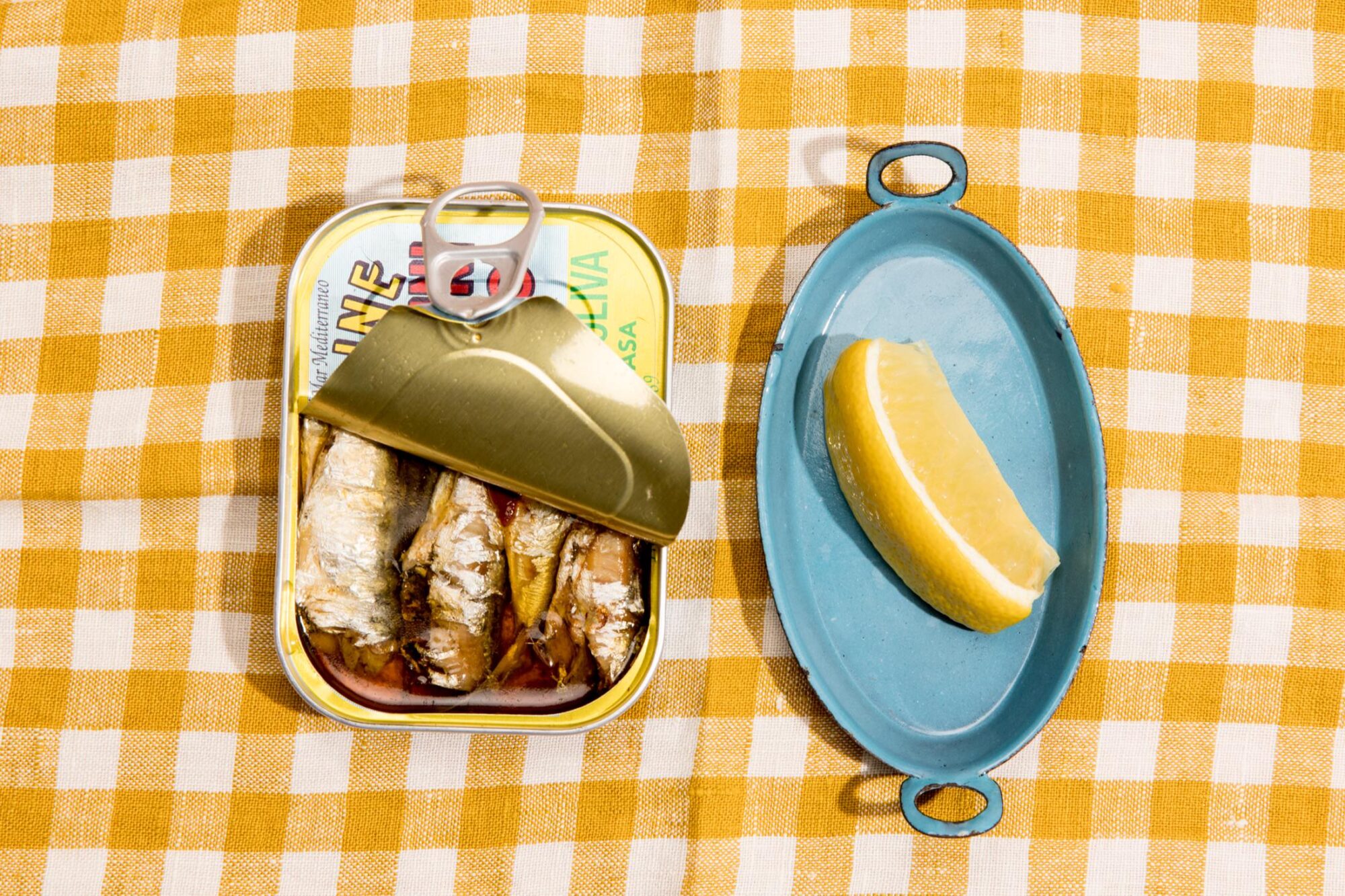 Seattle's best commercial food and hospitality photographer An opened tin of sardines and a blue oval plate with a lemon wedge sit on a yellow and white checkered tablecloth, perfectly styled by Seattles best commercial food lifestyle photographer. captured by seattle's best food and lifestyle photographer Brooke Fitts