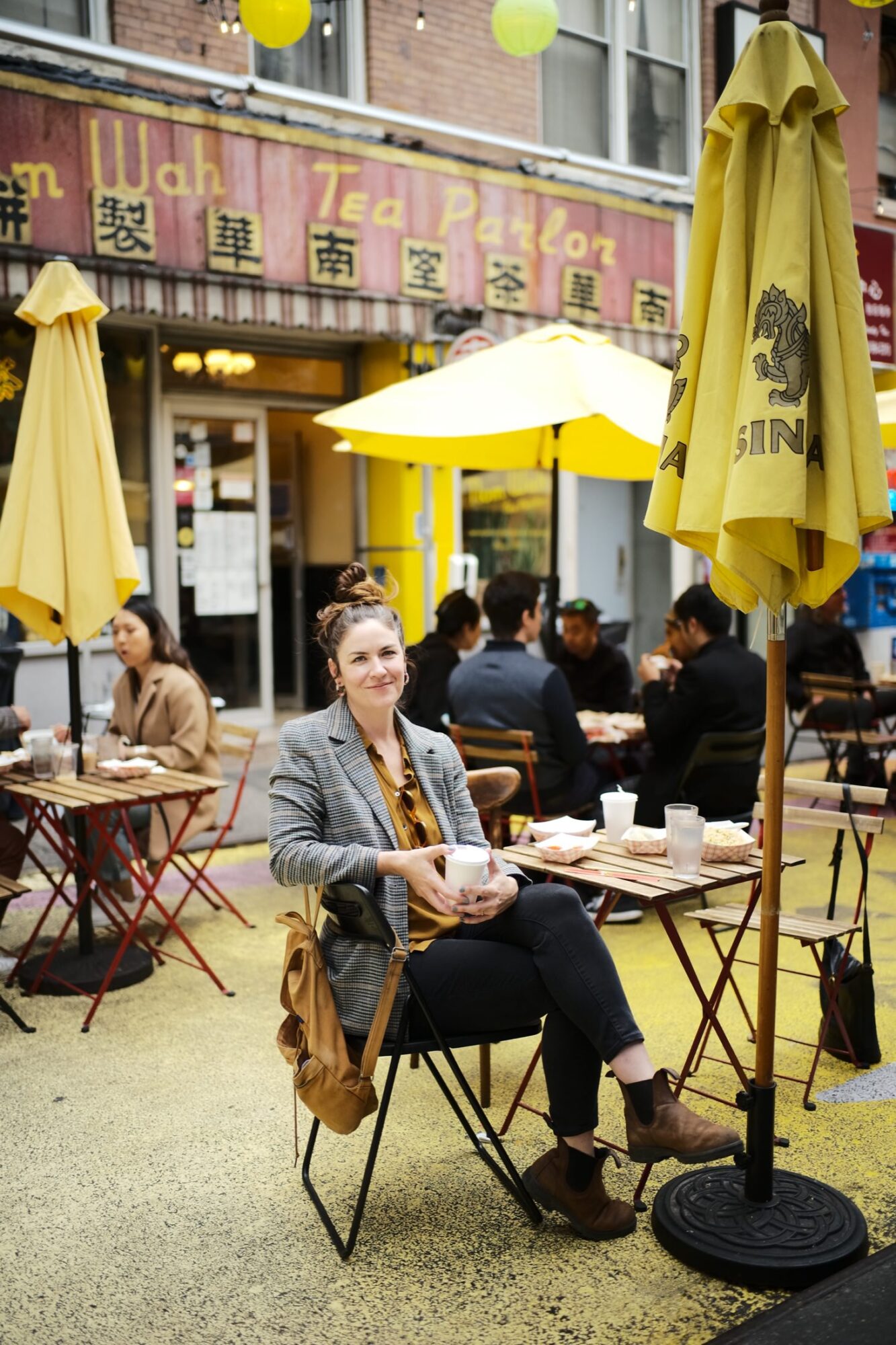 Seattle's best commercial food and hospitality photographer A woman sits at an outdoor café table, smiling at the camera with a cup in hand. Captured by Seattles best commercial food lifestyle photographer, the lively city street features yellow umbrellas and a Tea Parlor sign in the background. captured by seattle's best food and lifestyle photographer Brooke Fitts
