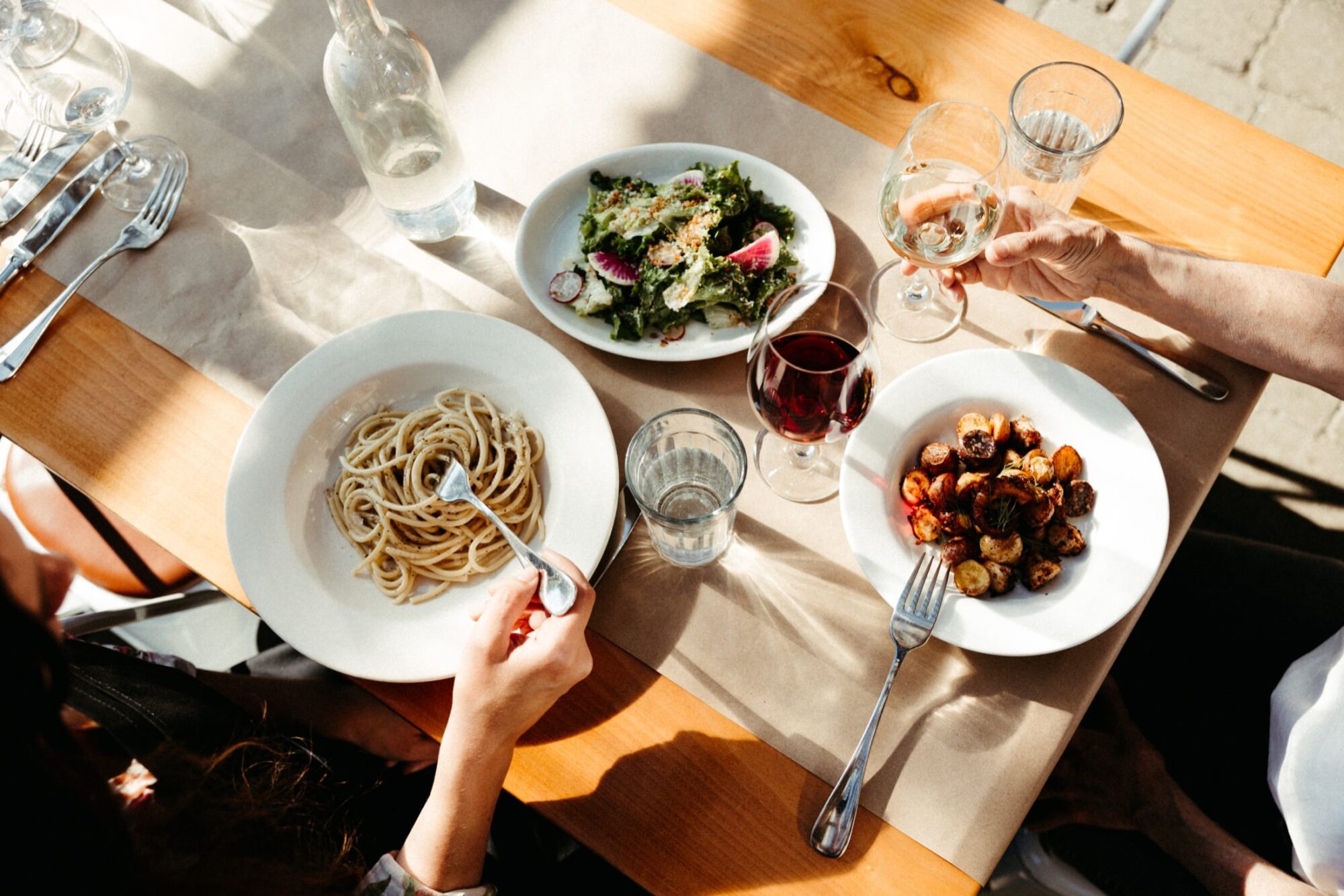 Seattle's best commercial food and hospitality photographer A table set for two with plates of pasta, salad, and roasted vegetables. Captured by Seattles best commercial food lifestyle photographer, sunlight streams over the wooden table as two diners toast with wine, creating a warm, inviting atmosphere. captured by seattle's best food and lifestyle photographer Brooke Fitts