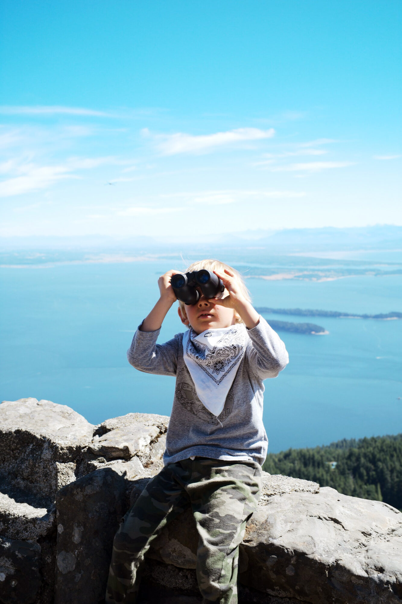 Seattle's best commercial food and hospitality photographer A young child wearing a bandana and camouflage pants sits on a rocky ledge, looking through binoculars. Captured by Seattles best commercial food lifestyle photographer, the scenic view reveals blue water, islands, and distant mountains under a clear sky. captured by seattle's best food and lifestyle photographer Brooke Fitts