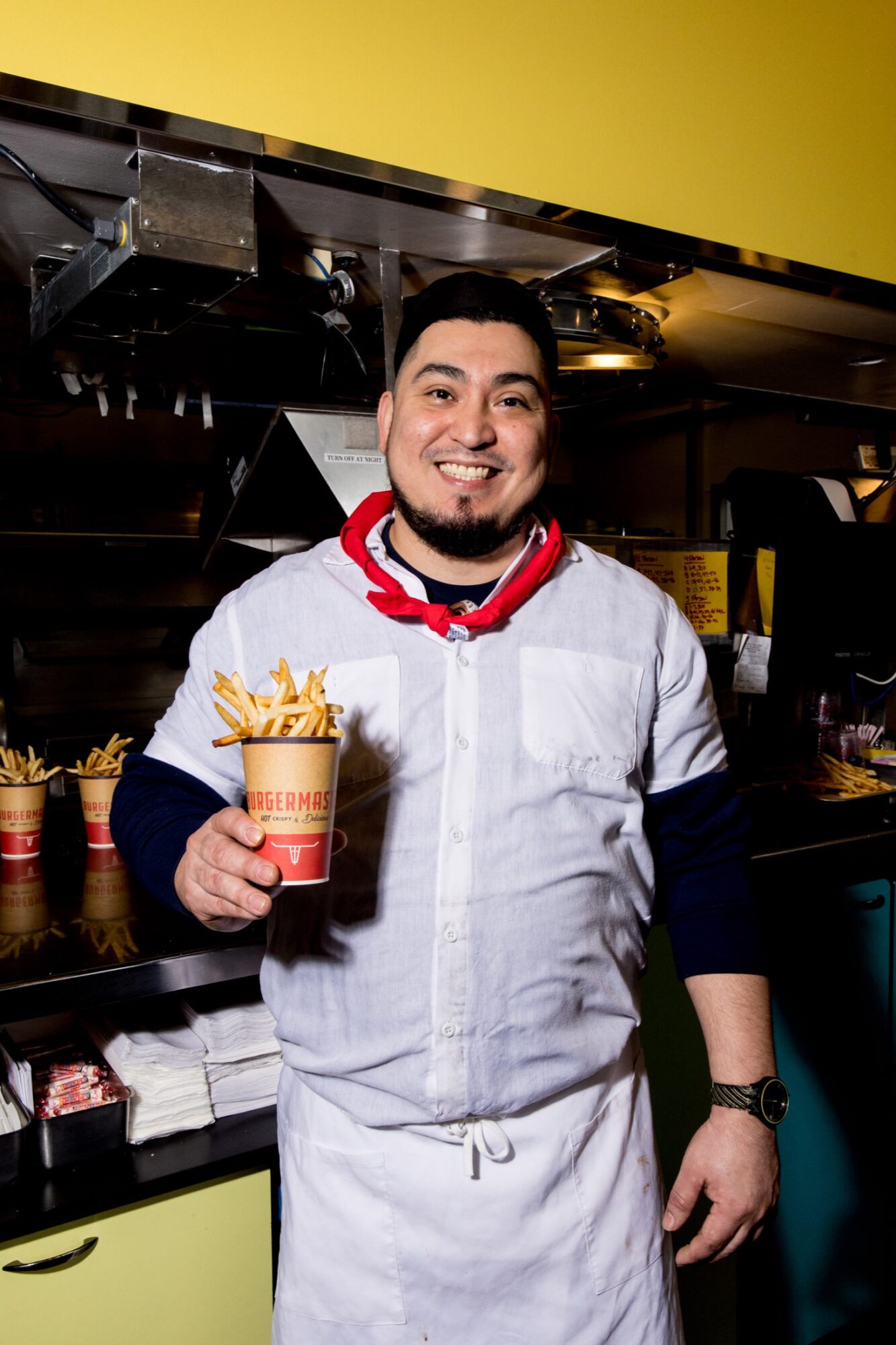 Seattle's best commercial food and hospitality photographer A smiling man in a white shirt and apron holds a cup of French fries in a restaurant kitchen, captured by Seattles best commercial food lifestyle photographer, with more fries and utensils visible in the background. captured by seattle's best food and lifestyle photographer Brooke Fitts