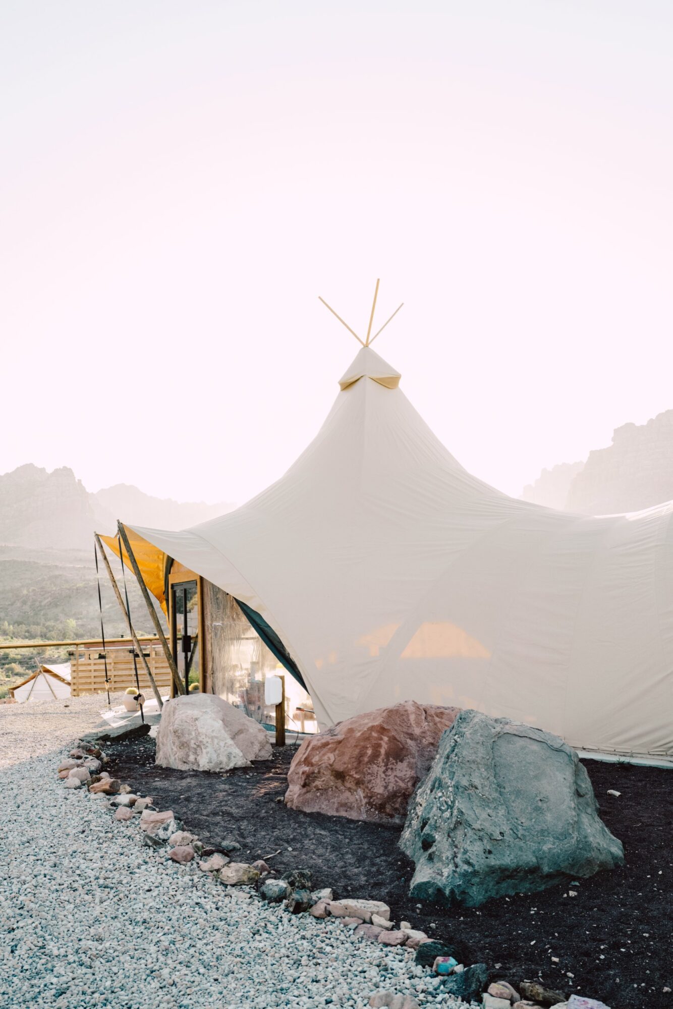 Seattle's best commercial food and hospitality photographer A large white glamping tent with a pointed roof stands on gravel near large rocks and a wooden fence, set against distant mountains under a bright, clear sky—captured by Seattles best commercial food lifestyle photographer. captured by seattle's best food and lifestyle photographer Brooke Fitts