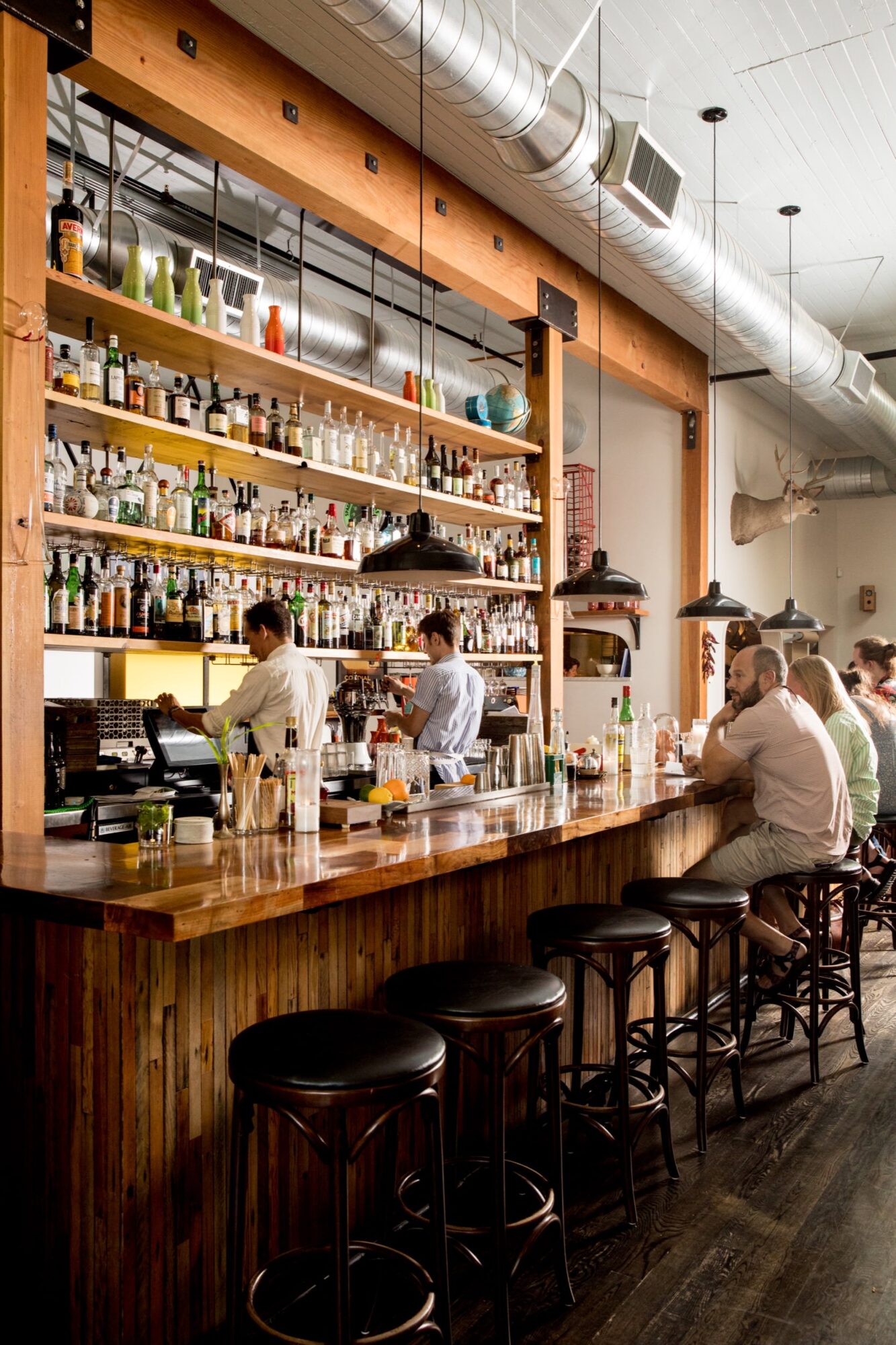 Seattle's best commercial food and hospitality photographer A cozy bar with wooden stools and a polished counter, lined with bottles and glasses. Captured by Seattles best commercial food lifestyle photographer, bartenders prepare drinks as patrons chat beneath industrial lights and shelves of liquor. captured by seattle's best food and lifestyle photographer Brooke Fitts