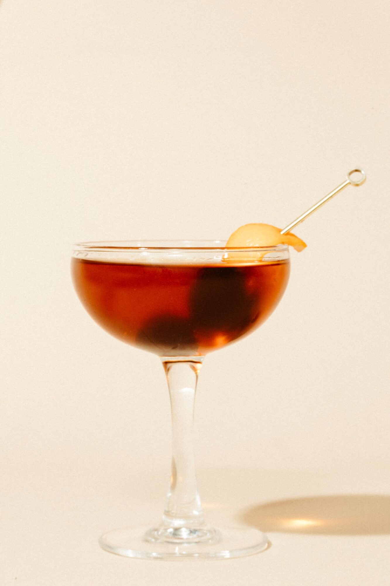 Seattle's best commercial food and hospitality photographer A coupe glass filled with a dark brown cocktail, garnished with a twist of orange peel on a cocktail pick, set against a plain beige background—captured by Seattles best commercial food lifestyle photographer. captured by seattle's best food and lifestyle photographer Brooke Fitts