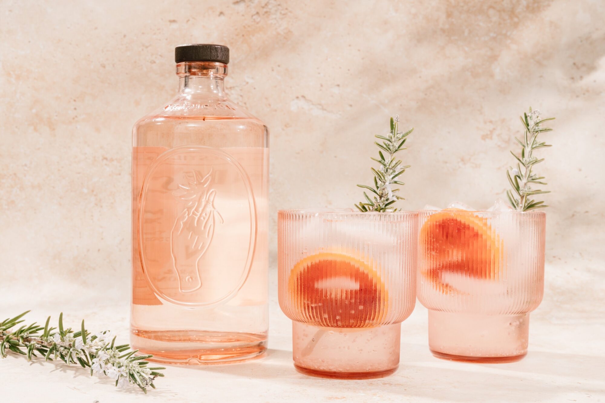 Seattle's best commercial food and hospitality photographer A pink bottle sits next to two glasses filled with ice, grapefruit slices, and rosemary sprigs; styled by Seattles best commercial food lifestyle photographer, all set against a neutral textured background. captured by seattle's best food and lifestyle photographer Brooke Fitts