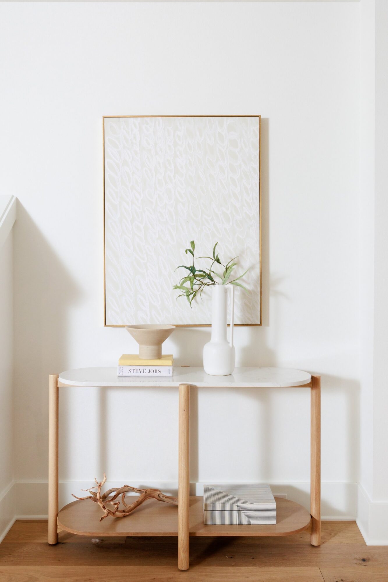 Seattle's best commercial food and hospitality photographer A minimalist console table with a light wood frame, styled by Seattles best commercial food lifestyle photographer, holds a beige vase with greenery, a white vase with a branch, stacked books, and driftwood. Neutral abstract art hangs above on the white wall. captured by seattle's best food and lifestyle photographer Brooke Fitts