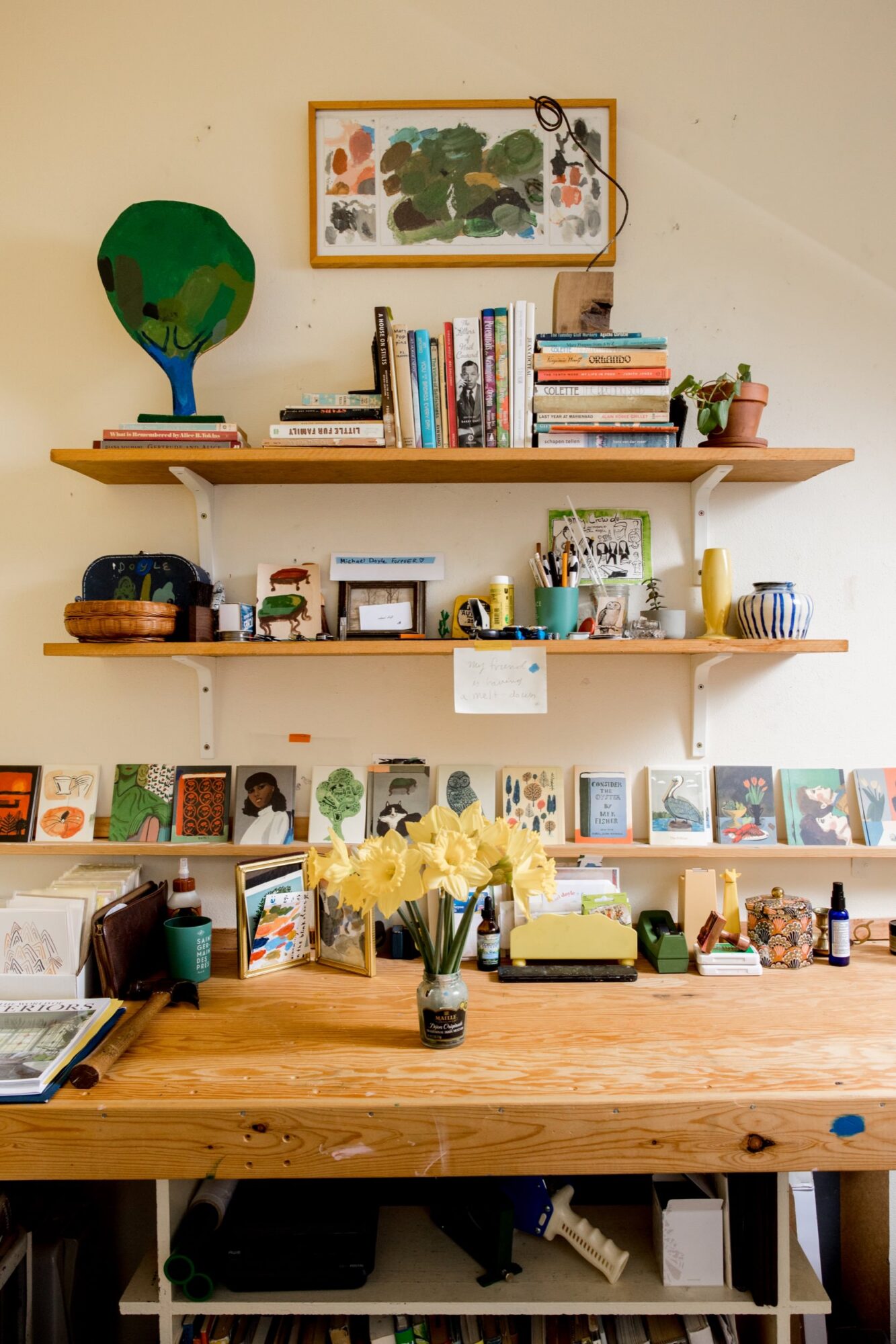 Seattle's best commercial food and hospitality photographer A cozy, organized desk with daffodils in a vase, shelves above holding books, art supplies, plants, and small art pieces sits against a cream wall decorated with colorful artwork—styled by Seattles best commercial food lifestyle photographer. captured by seattle's best food and lifestyle photographer Brooke Fitts