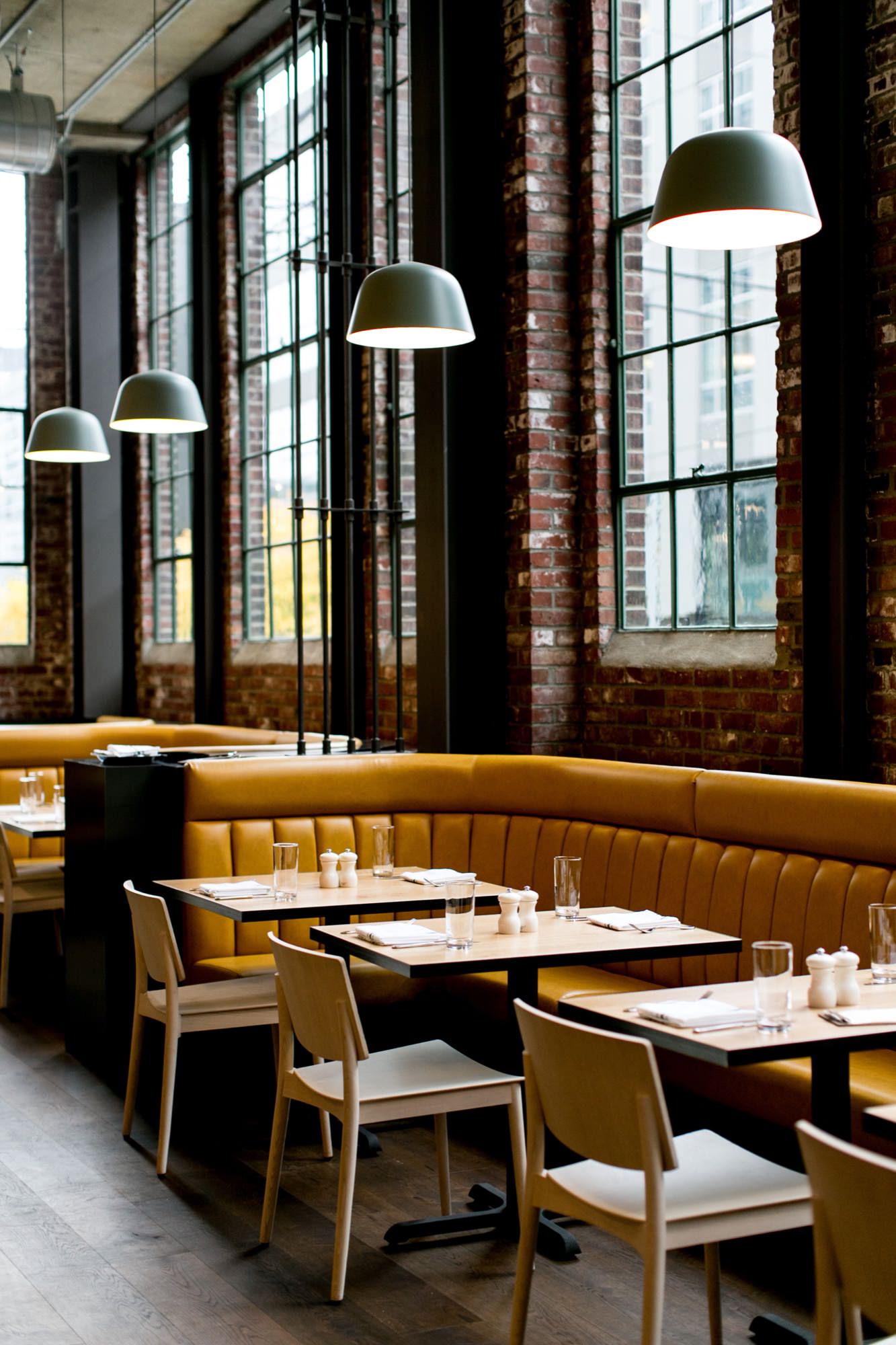 Seattle's best commercial food and hospitality photographer A modern restaurant captured by Seattles best commercial food lifestyle photographer, featuring tall windows, exposed brick walls, mustard yellow booth seating, white chairs, and pendant lights set over dark wooden tables. captured by seattle's best food and lifestyle photographer Brooke Fitts