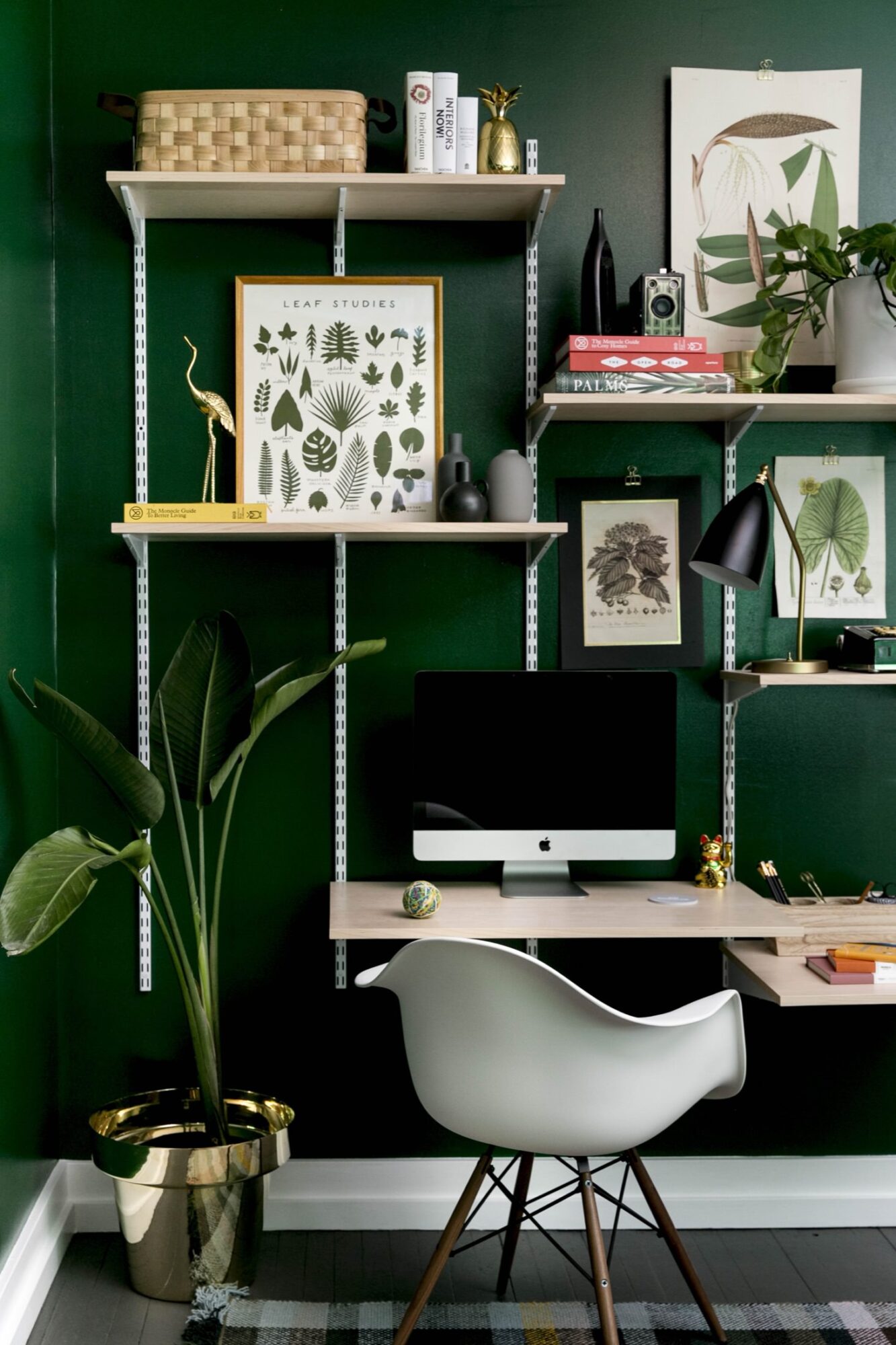 Seattle's best commercial food and hospitality photographer A modern home office with a dark green wall, floating shelves with plants, books, leaf art prints, and gold decor—styled like Seattles best commercial food lifestyle photographer arranged it. An iMac sits on a wooden desk; a white chair and large potted plant complete the look. captured by seattle's best food and lifestyle photographer Brooke Fitts