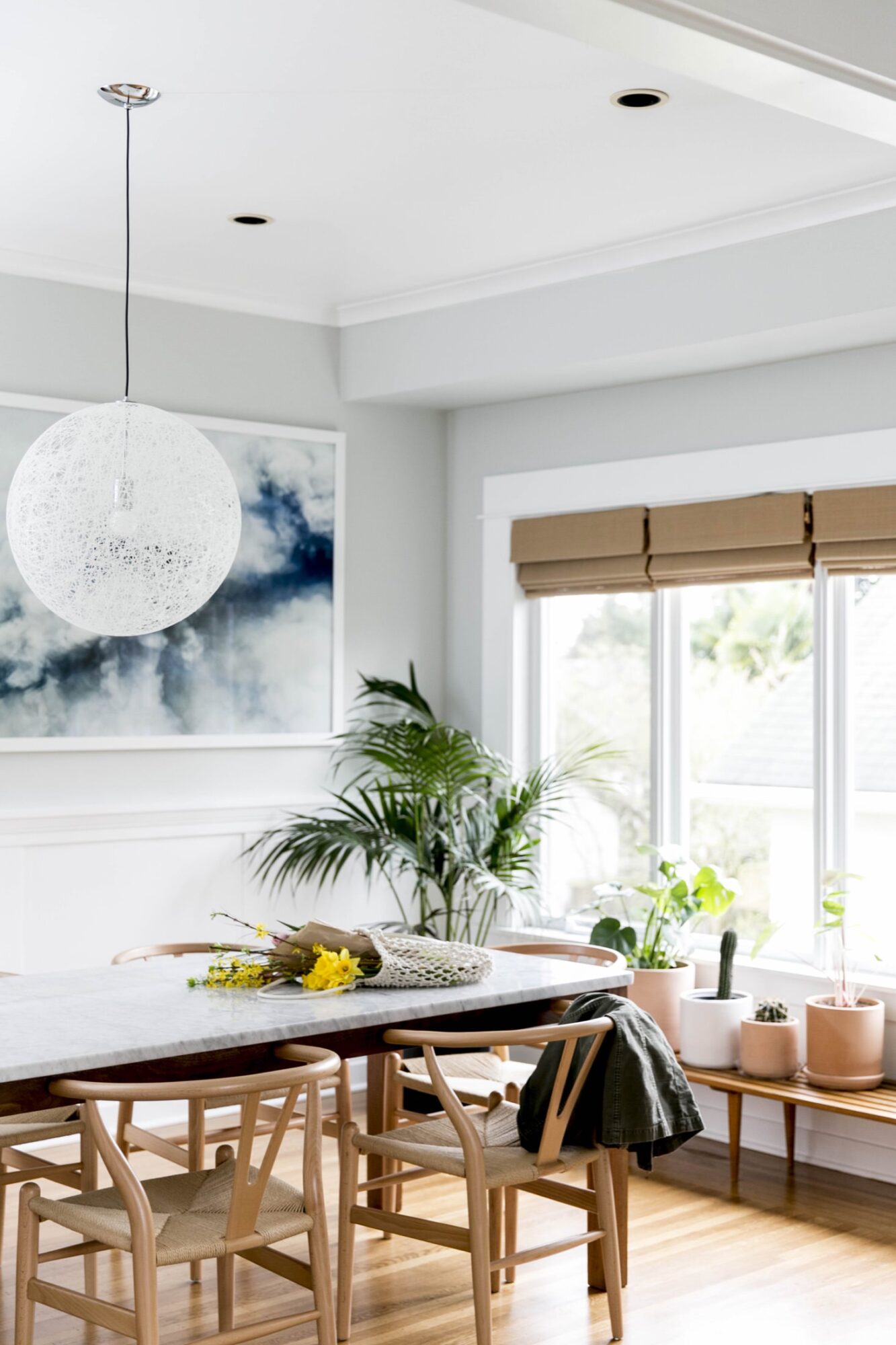 Seattle's best commercial food and hospitality photographer Bright dining room with large windows, wooden table and chairs, potted plants, a modern white pendant light, and a framed abstract painting—captured by Seattles best commercial food lifestyle photographer as sunlight streams in through tan window blinds. captured by seattle's best food and lifestyle photographer Brooke Fitts