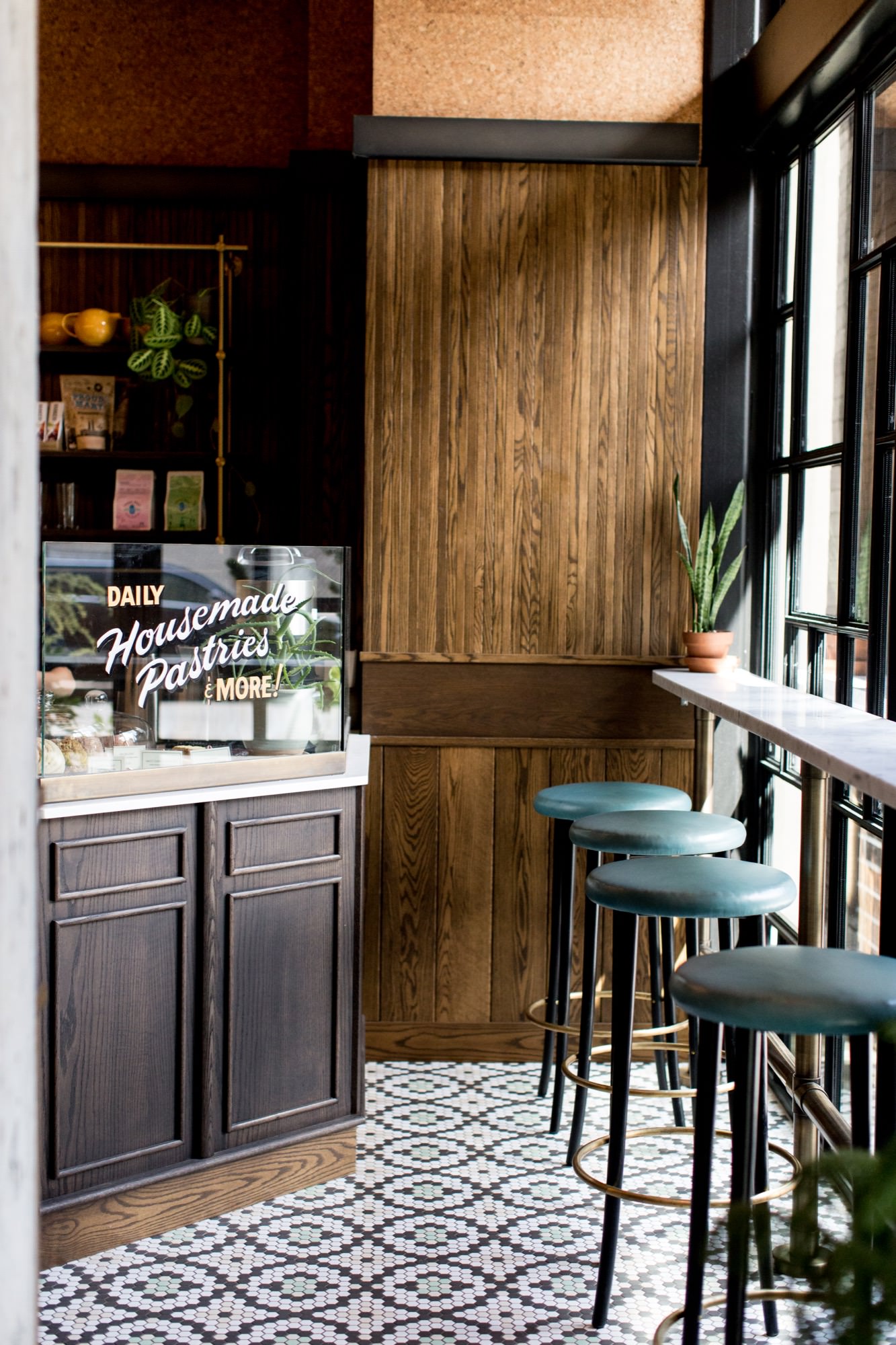 Seattle's best commercial interiors and hospitality photographer A cozy café corner, beautifully captured by a Seattle commercial interior design photographer for restaurants and hotels, features dark wood cabinets, a pastry display case, marble counter with high stools, patterned tile floor, and a potted plant on the windowsill. captured by seattle's best food and lifestyle photographer Brooke Fitts