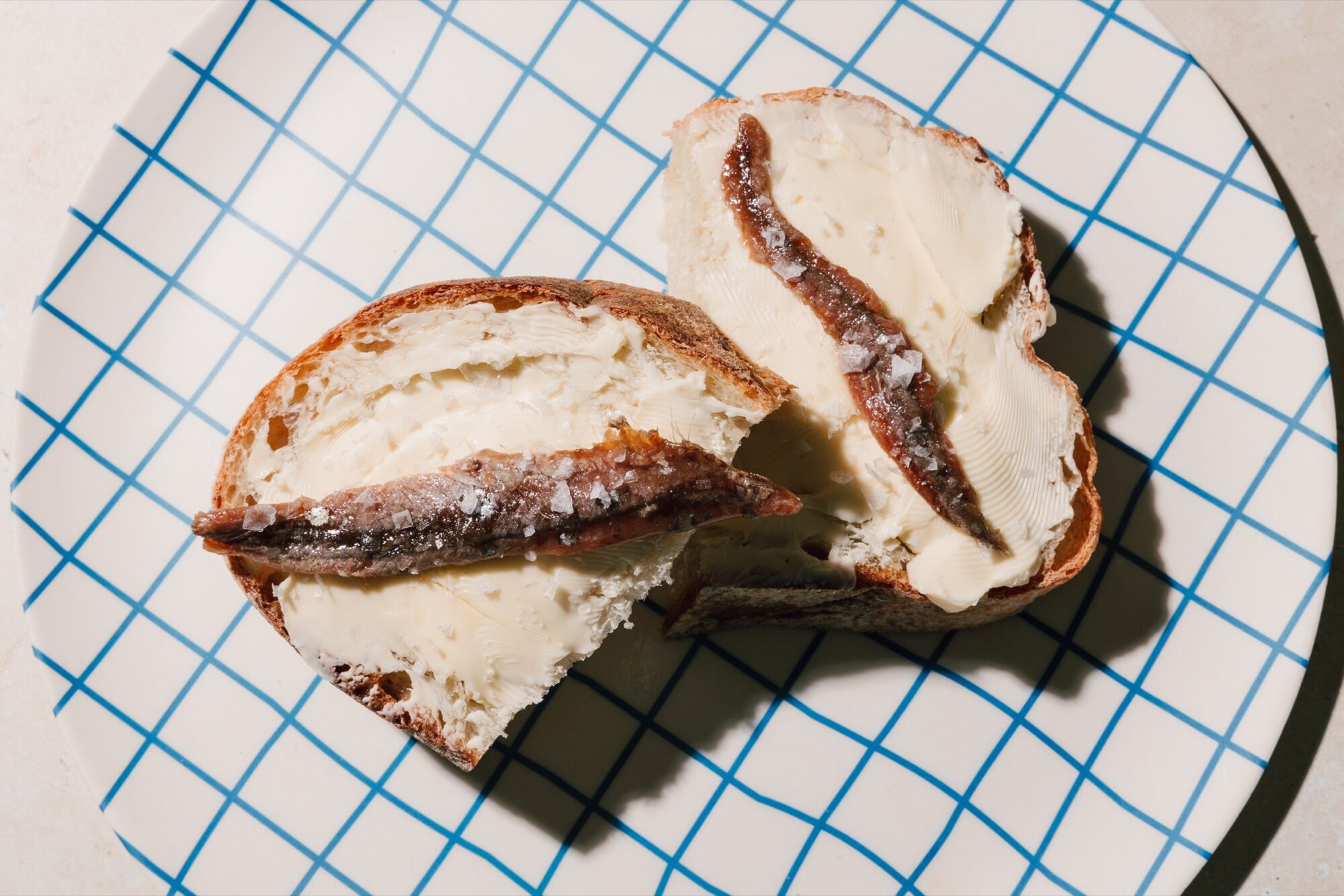 Seattle's best commercial food and hospitality photographer Two slices of bread with butter and anchovy fillets are placed on a white plate with a blue grid pattern, captured by Seattles best commercial food lifestyle photographer. captured by seattle's best food and lifestyle photographer Brooke Fitts