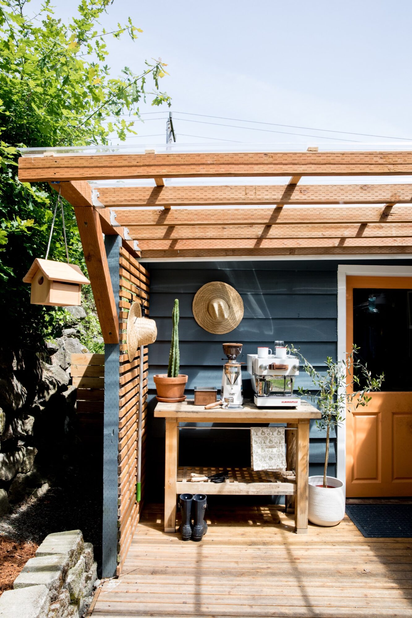Seattle's best commercial food and hospitality photographer A cozy outdoor coffee bar setup under a wooden pergola, captured by Seattles best commercial food lifestyle photographer, features a table with a coffee machine, potted plants, hats on the wall, a hanging birdhouse, and an orange door into a blue house. captured by seattle's best food and lifestyle photographer Brooke Fitts
