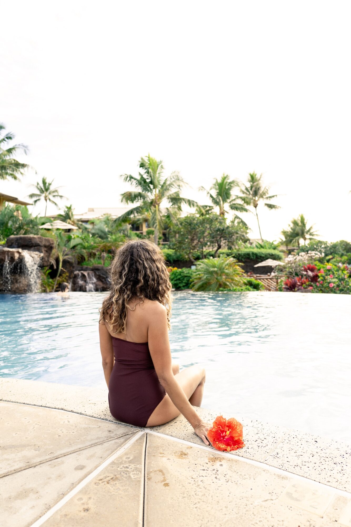 Seattle's best commercial food and hospitality photographer A woman with curly hair in a maroon swimsuit sits at the edge of a pool holding an orange flower, surrounded by palm trees and tropical plants—captured by Seattles best commercial food lifestyle photographer. captured by seattle's best food and lifestyle photographer Brooke Fitts