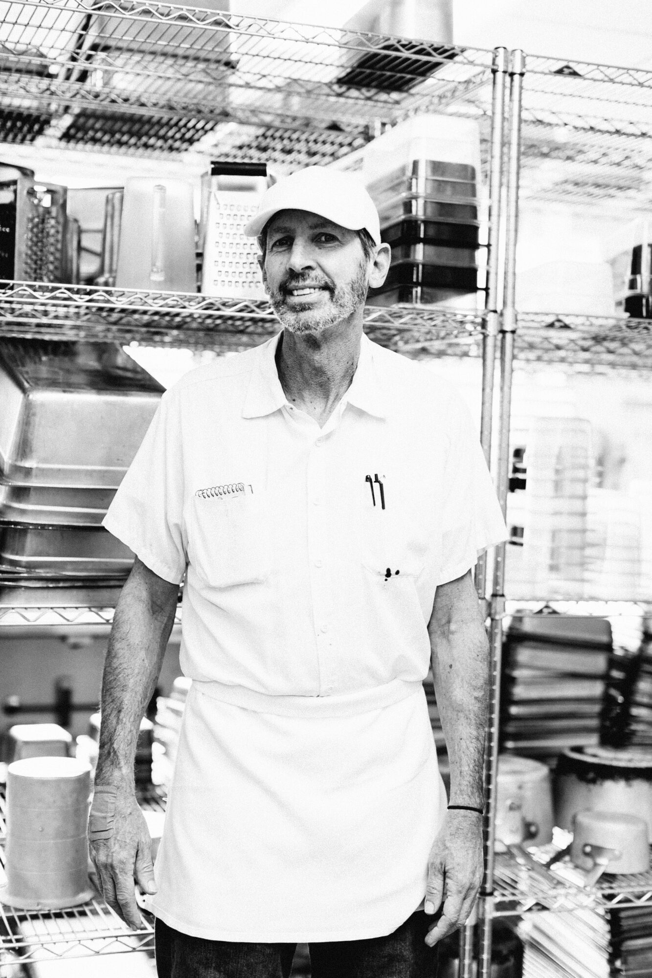 Seattle's best commercial food and hospitality photographer A man wearing a white shirt, apron, and cap stands in front of metal shelves filled with kitchen equipment in a commercial kitchen, captured in black and white by Seattles best commercial food lifestyle photographer. captured by seattle's best food and lifestyle photographer Brooke Fitts