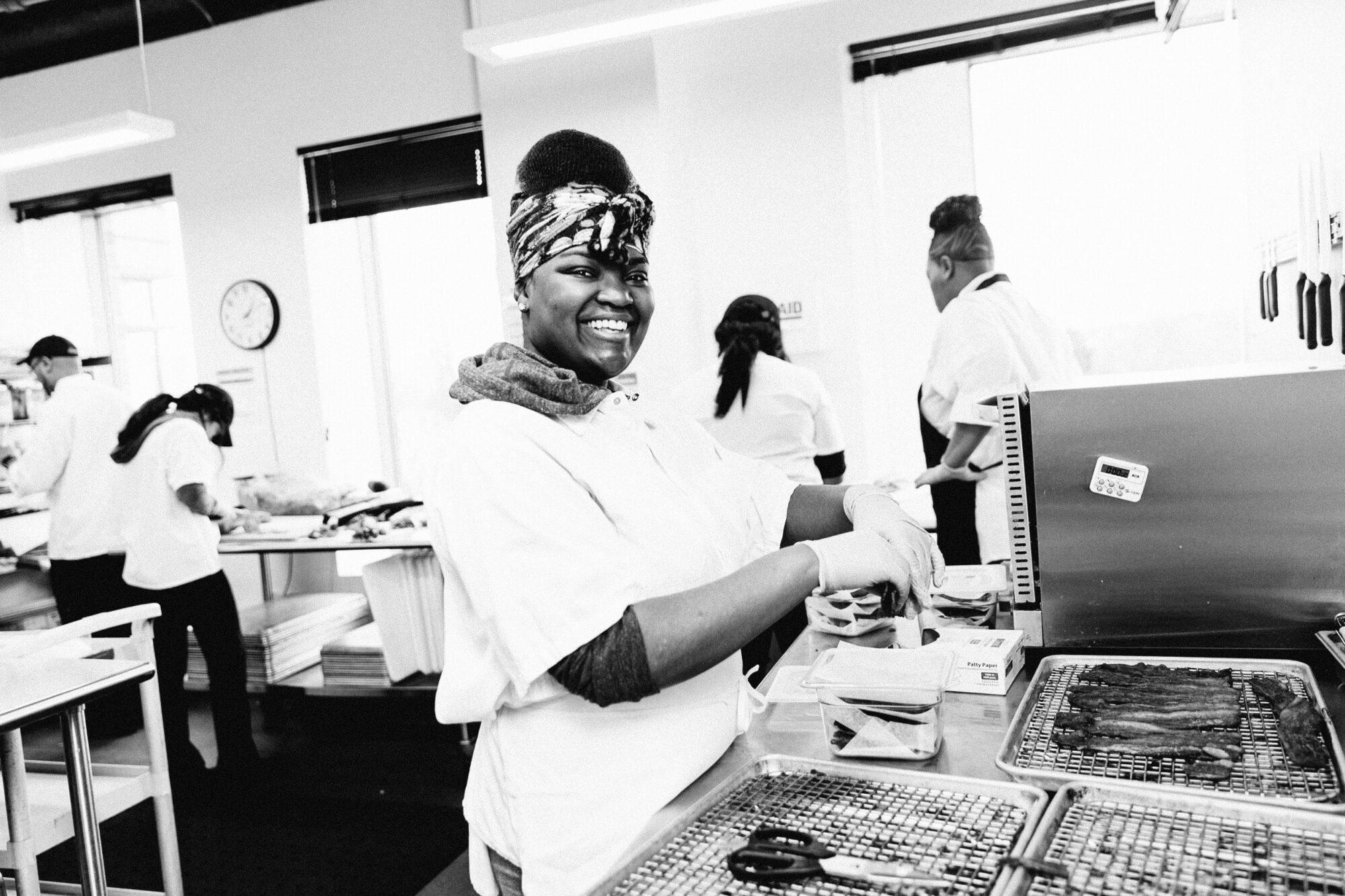 Seattle's best commercial food and hospitality photographer A woman in a headscarf smiles while preparing food in a bright, busy kitchen—captured by Seattles best commercial food lifestyle photographer. Baking trays and packaged ingredients are spread out on the counter before her. captured by seattle's best food and lifestyle photographer Brooke Fitts