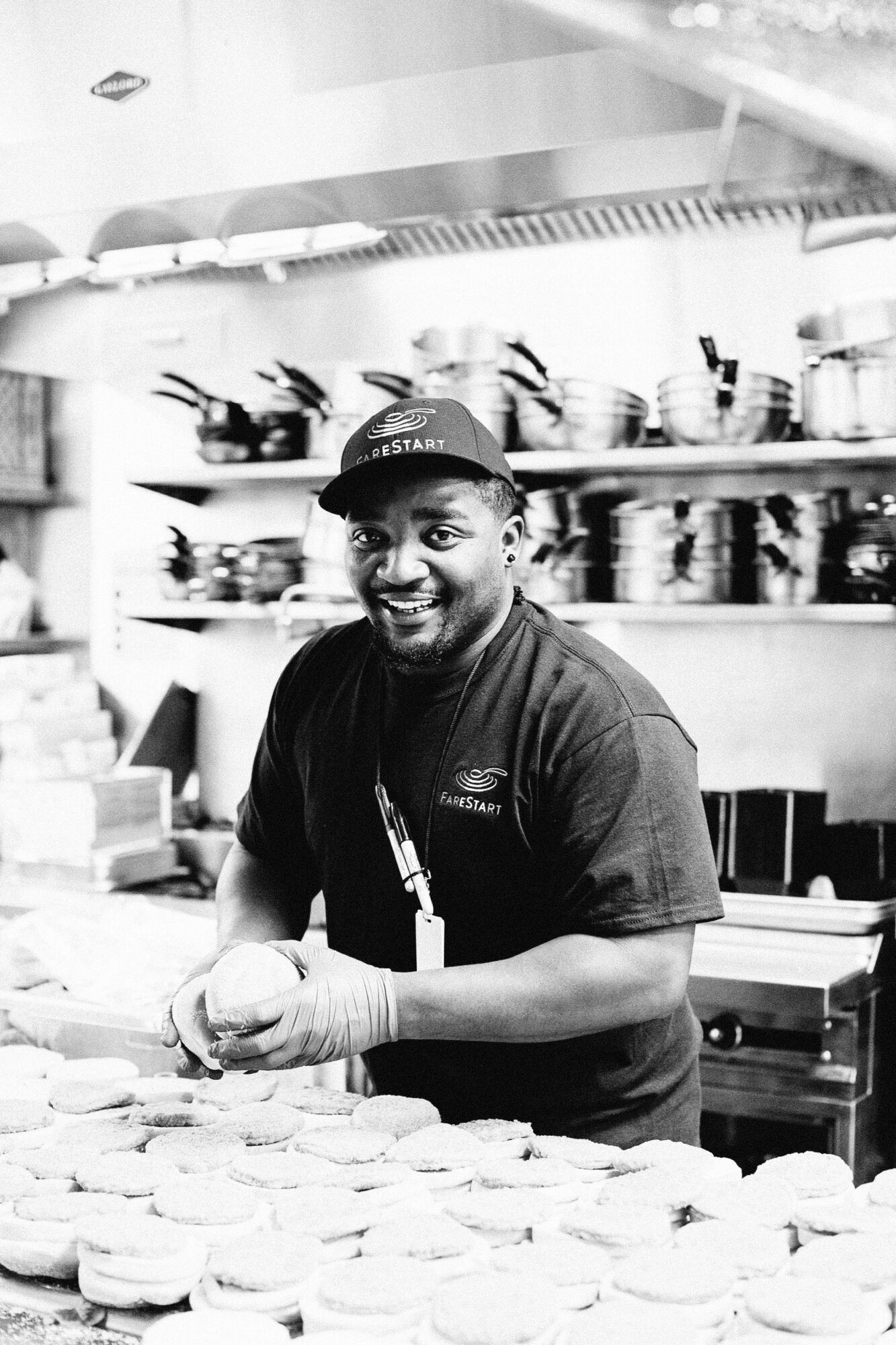 Seattle's best commercial food and hospitality photographer A smiling man in a cap and gloves prepares sandwiches in a commercial kitchen, standing behind a counter with many sandwiches—capturing the scene as Seattles best commercial food lifestyle photographer would. Pots and utensils line the shelves behind him. captured by seattle's best food and lifestyle photographer Brooke Fitts
