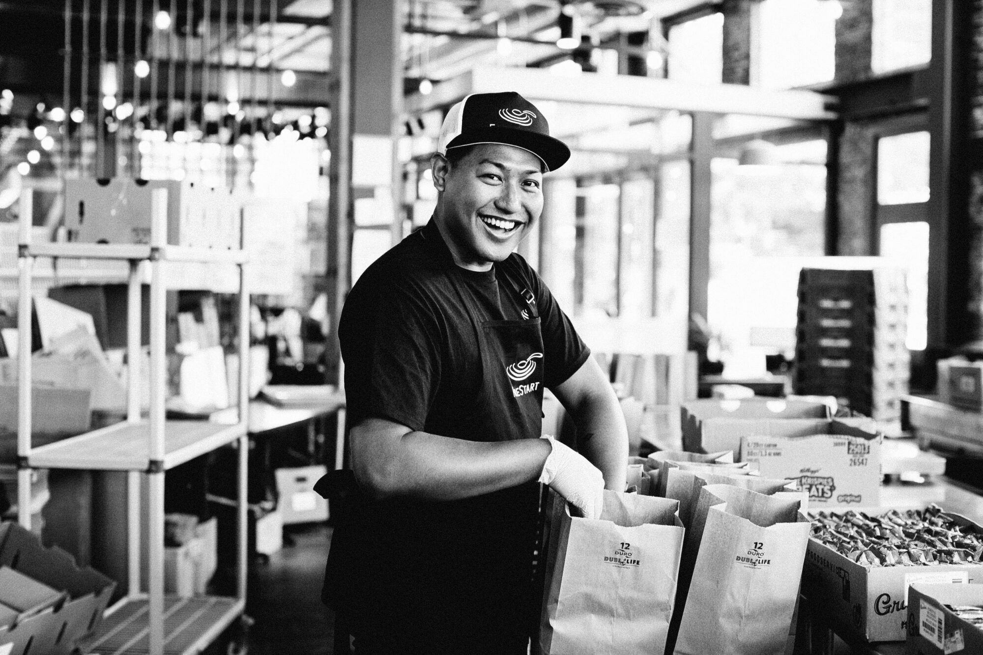Seattle's best commercial food and hospitality photographer A smiling worker, captured by Seattles best commercial food lifestyle photographer, wears a cap and gloves while filling paper bags with groceries inside a bright, modern store. The black and white image highlights shelves and boxes in the background. captured by seattle's best food and lifestyle photographer Brooke Fitts