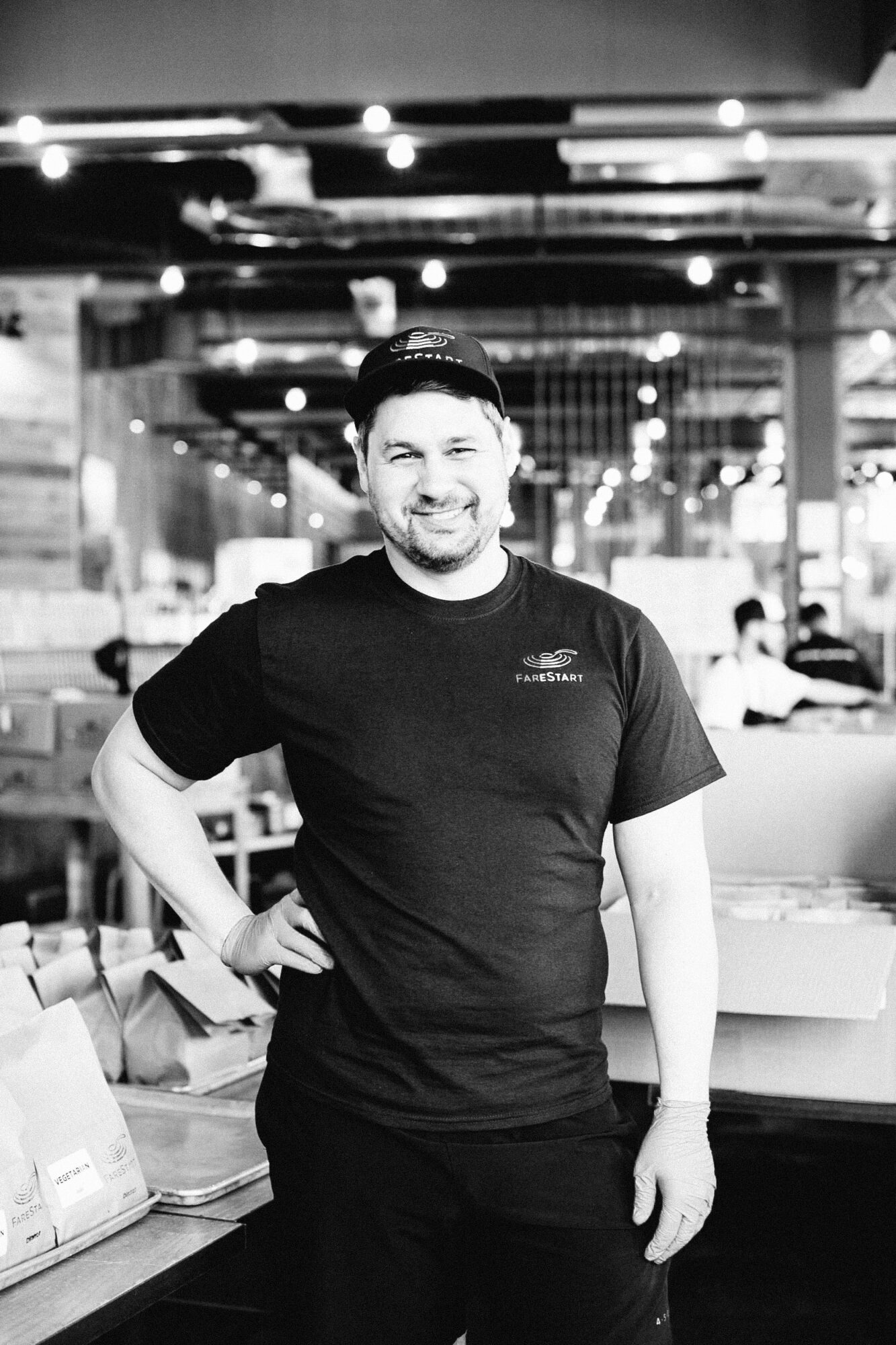 Seattle's best commercial food and hospitality photographer A man in a dark t-shirt and cap stands smiling in a modern, busy restaurant or café, perfectly capturing the atmosphere as Seattles best commercial food lifestyle photographer would. Paper bags and other staff are visible in the background. captured by seattle's best food and lifestyle photographer Brooke Fitts