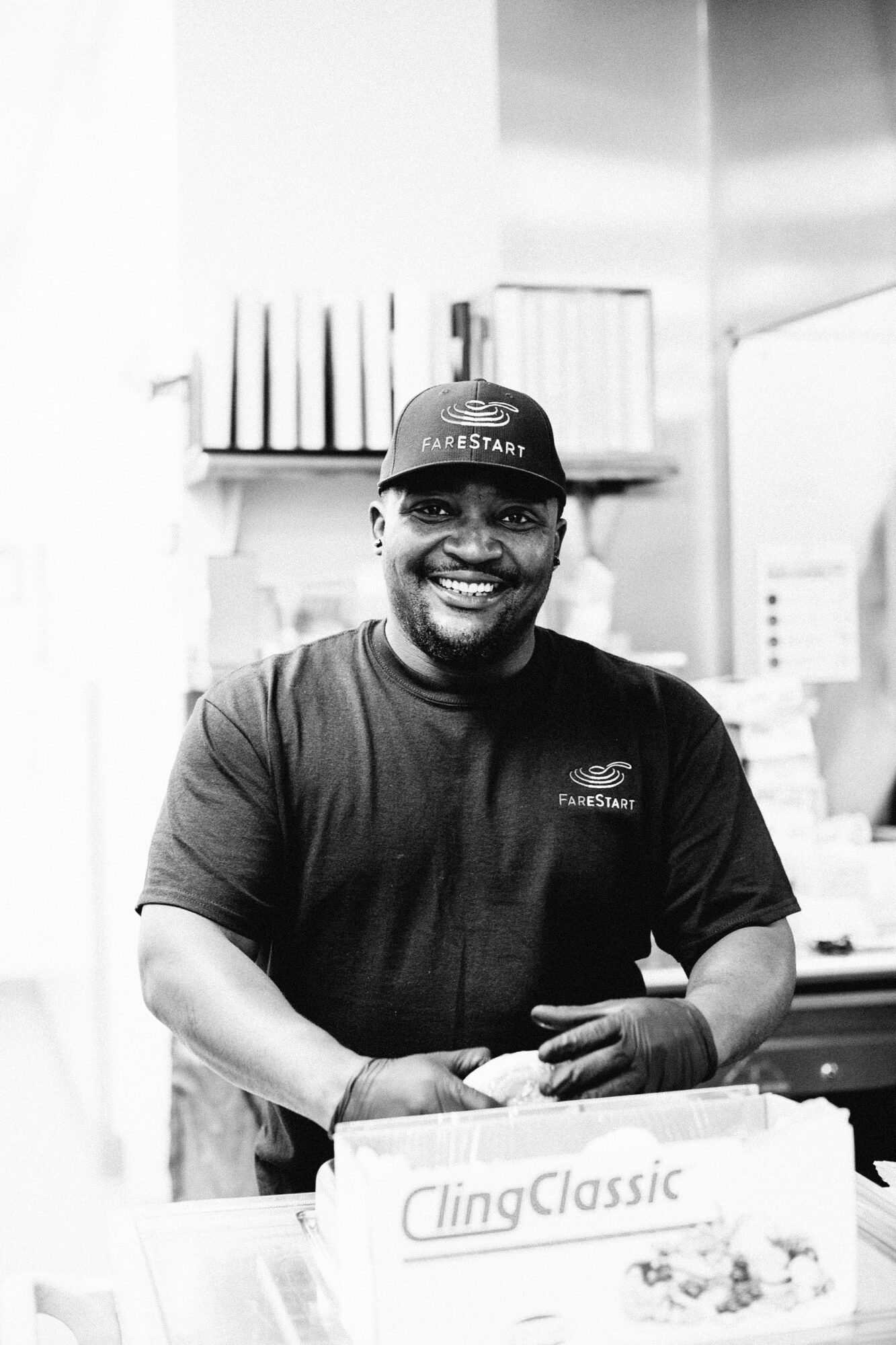 Seattle's best commercial food and hospitality photographer A smiling man wearing a FareStart cap and t-shirt stands in a kitchen, handling food with gloved hands near a box labeled Cling Classic—captured by Seattles best commercial food lifestyle photographer. Shelves with supplies are visible in the background. captured by seattle's best food and lifestyle photographer Brooke Fitts