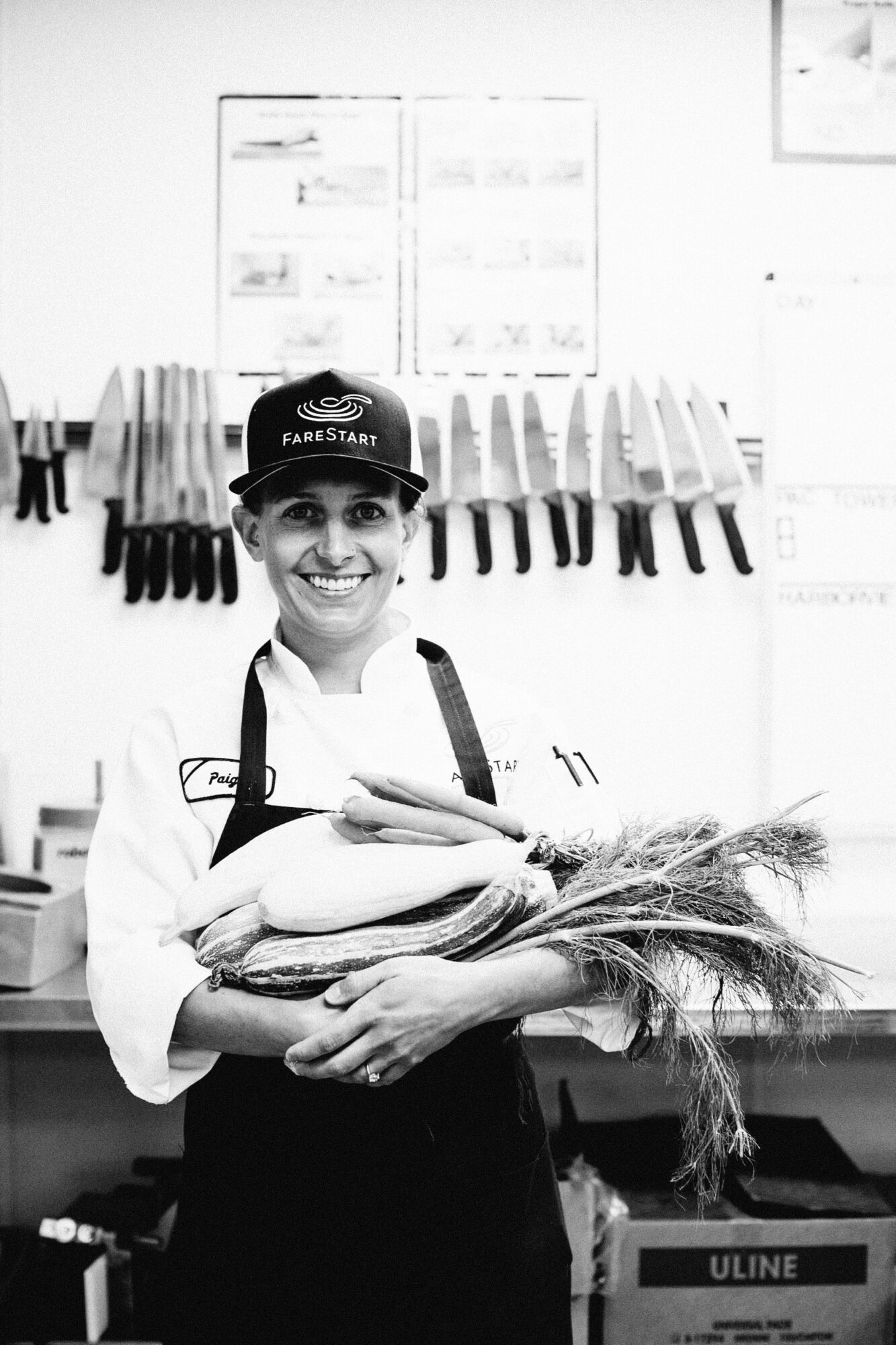 Seattle's best commercial food and hospitality photographer A smiling chef in uniform and a FareStart cap stands in a kitchen, holding fresh carrots and leeks. Knives are neatly arranged on the wall behind them in this black and white image by Seattles best commercial food lifestyle photographer. captured by seattle's best food and lifestyle photographer Brooke Fitts
