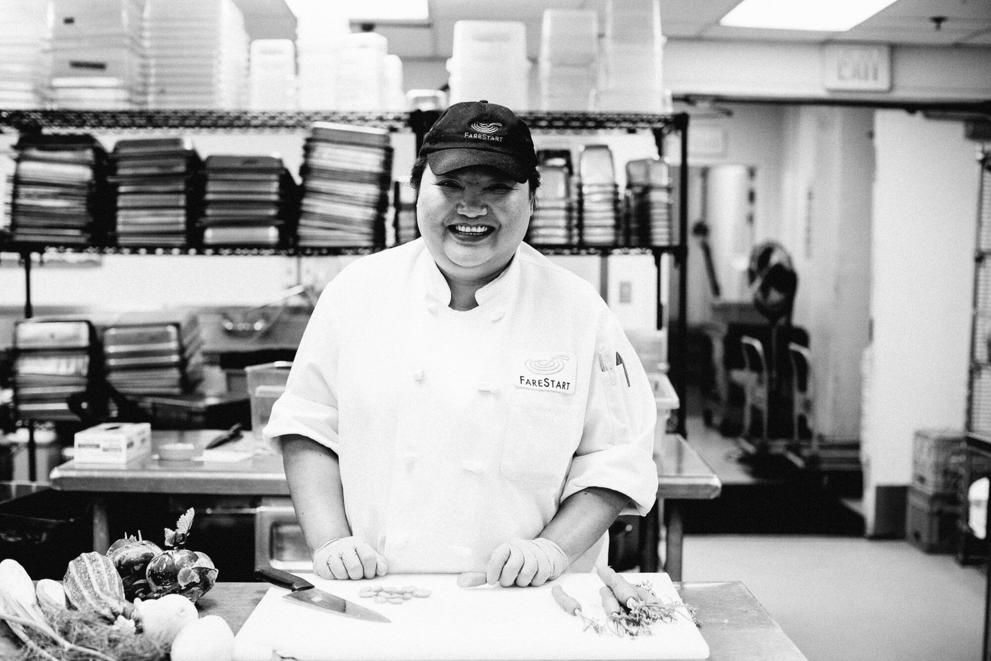 Seattle's best commercial food and hospitality photographer A chef in a uniform and cap stands smiling behind a counter in a commercial kitchen, with chopped vegetables and a knife on a cutting board—captured by Seattles best commercial food lifestyle photographer. Shelves with containers fill the background. captured by seattle's best food and lifestyle photographer Brooke Fitts
