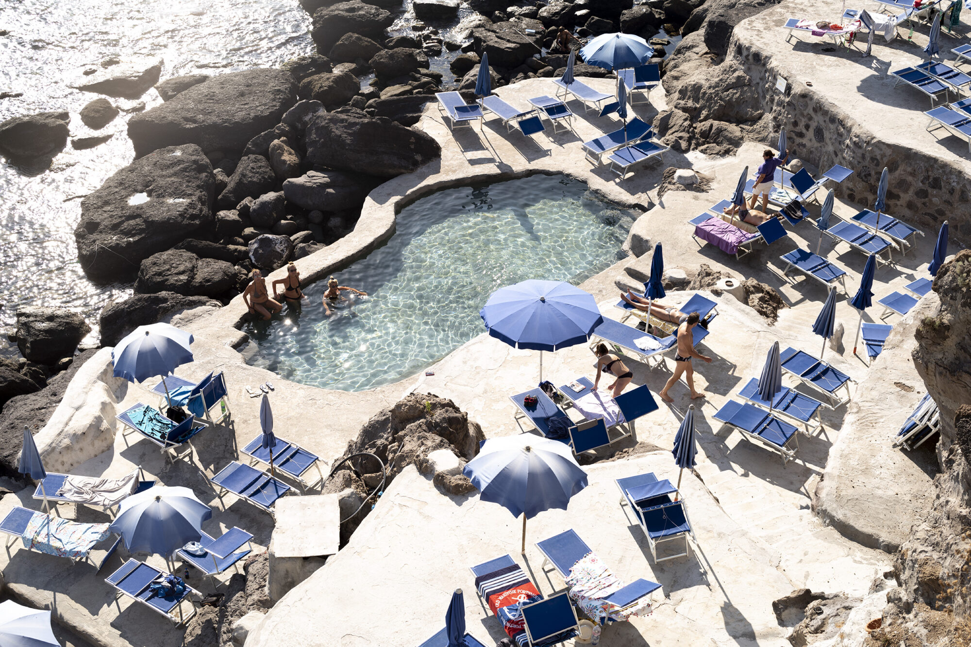 Seattle's best commercial food and hospitality photographer People relax by a small, rocky seaside pool surrounded by blue loungers and umbrellas. Captured with the artistry of Seattles best commercial food lifestyle photographer, sunlight reflects off the water for a serene Mediterranean atmosphere. captured by seattle's best food and lifestyle photographer Brooke Fitts