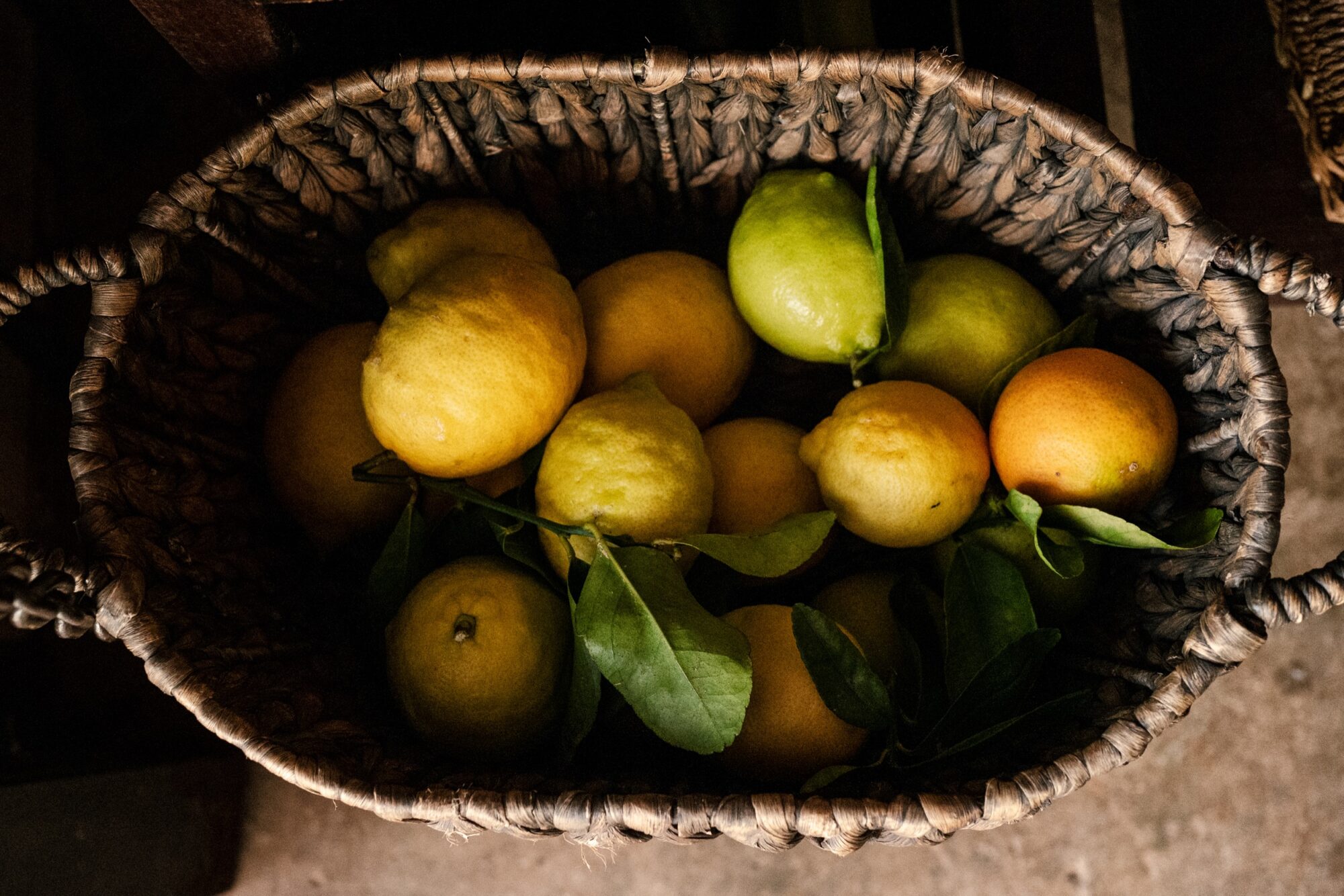 Seattle's best commercial food and hospitality photographer A woven basket filled with yellow lemons, a few green and orange citrus fruits, and green leaves, artfully styled on a rustic surface by Seattles best commercial food lifestyle photographer. captured by seattle's best food and lifestyle photographer Brooke Fitts