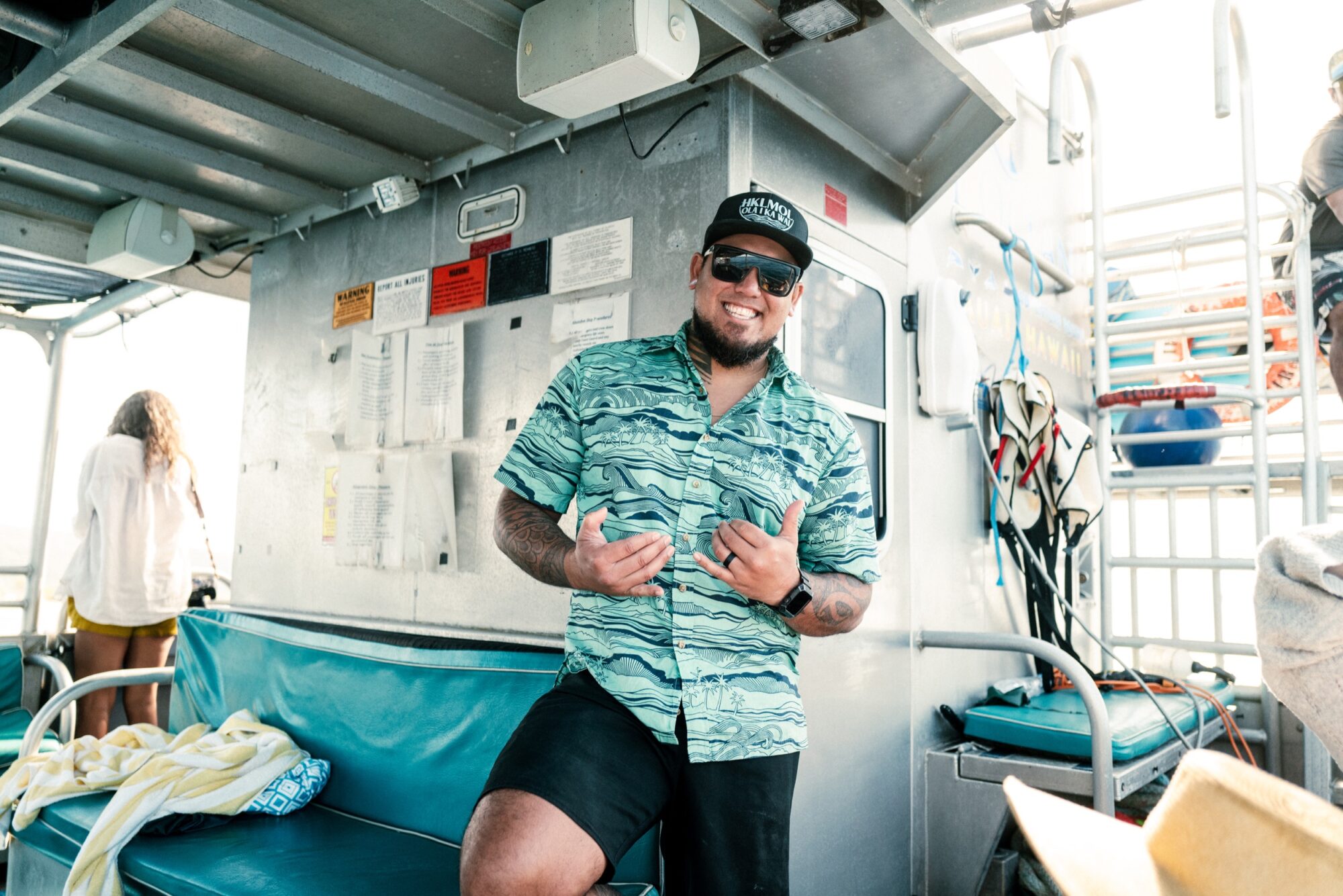 Seattle's best commercial food and hospitality photographer A man wearing sunglasses, a patterned shirt, and a hat smiles at the camera and gestures with both hands while standing on a boat—captured by Seattles best commercial food lifestyle photographer—with seating, equipment, and posted notices behind him. captured by seattle's best food and lifestyle photographer Brooke Fitts