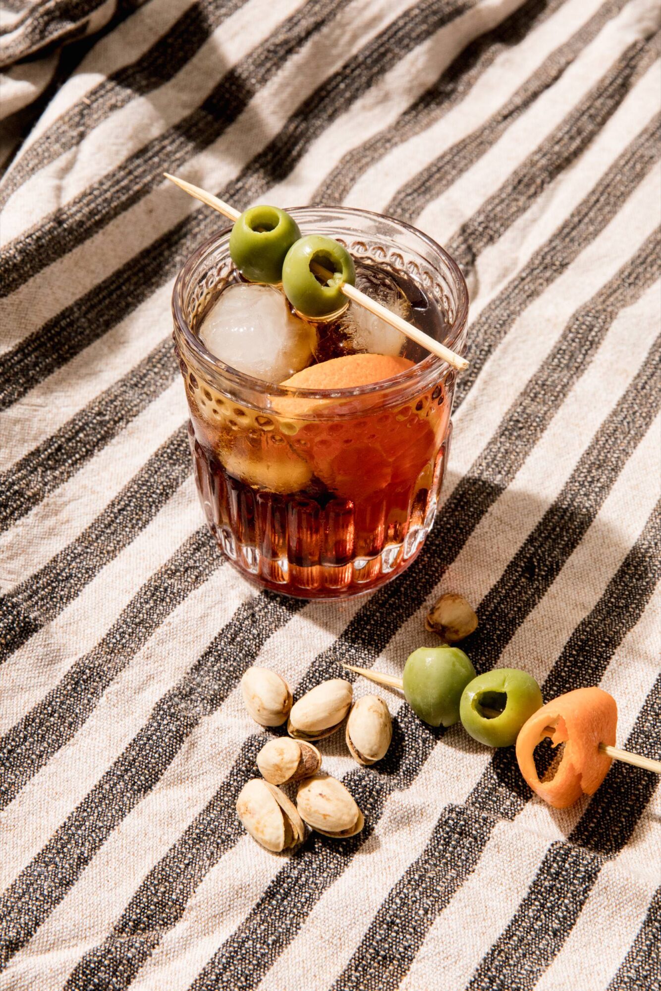 Seattle's best commercial food and hospitality photographer A glass of iced cocktail garnished with green olives on a skewer sits on a striped fabric, skillfully captured by Seattle’s best commercial food lifestyle photographer, with pistachios, an olive, and a carrot slice scattered nearby. captured by seattle's best food and lifestyle photographer Brooke Fitts